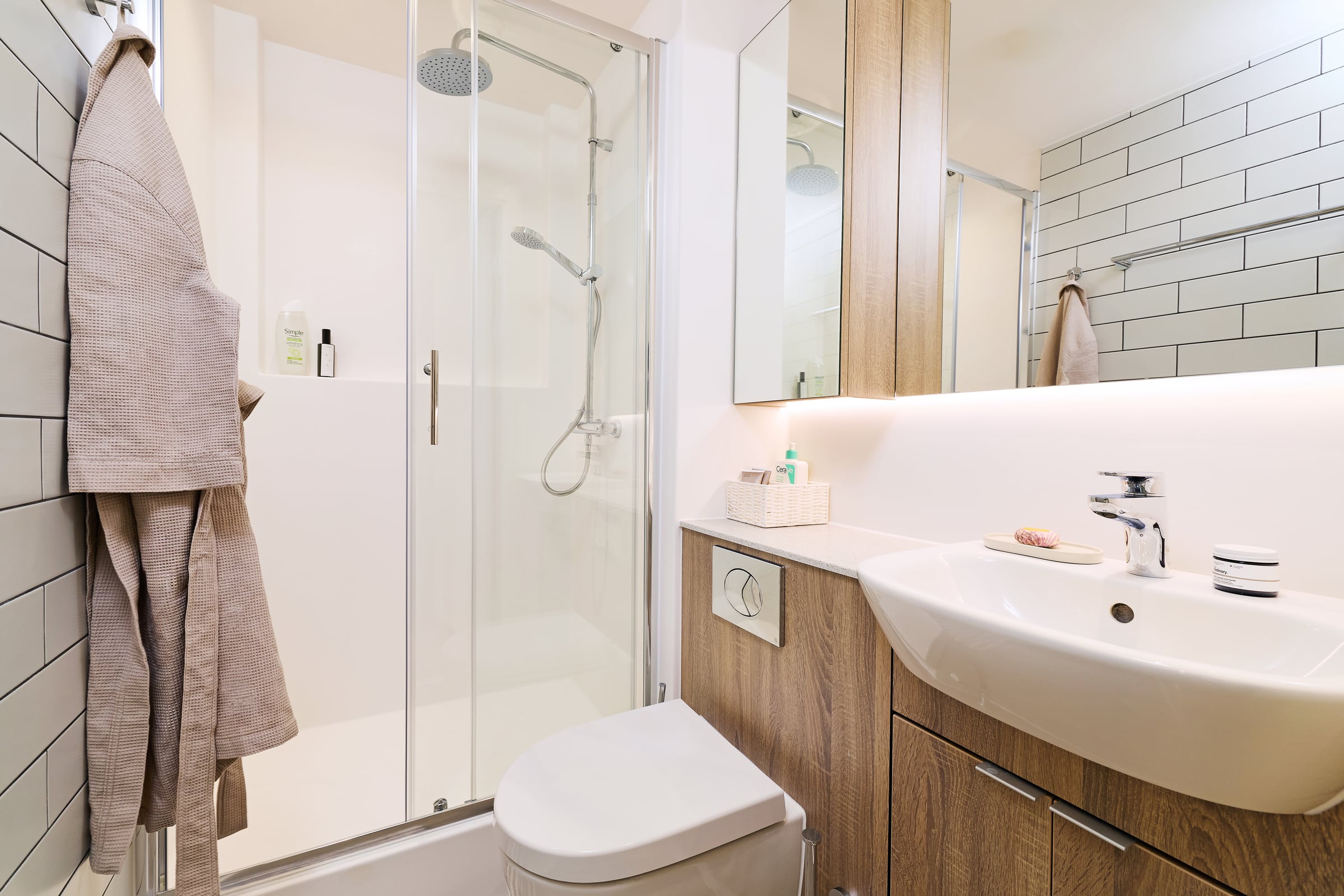 Modern bathroom with a glass shower, wooden cabinets, a white sink, and a toilet. A beige robe hangs on the wall and toiletries are neatly arranged on the countertop.