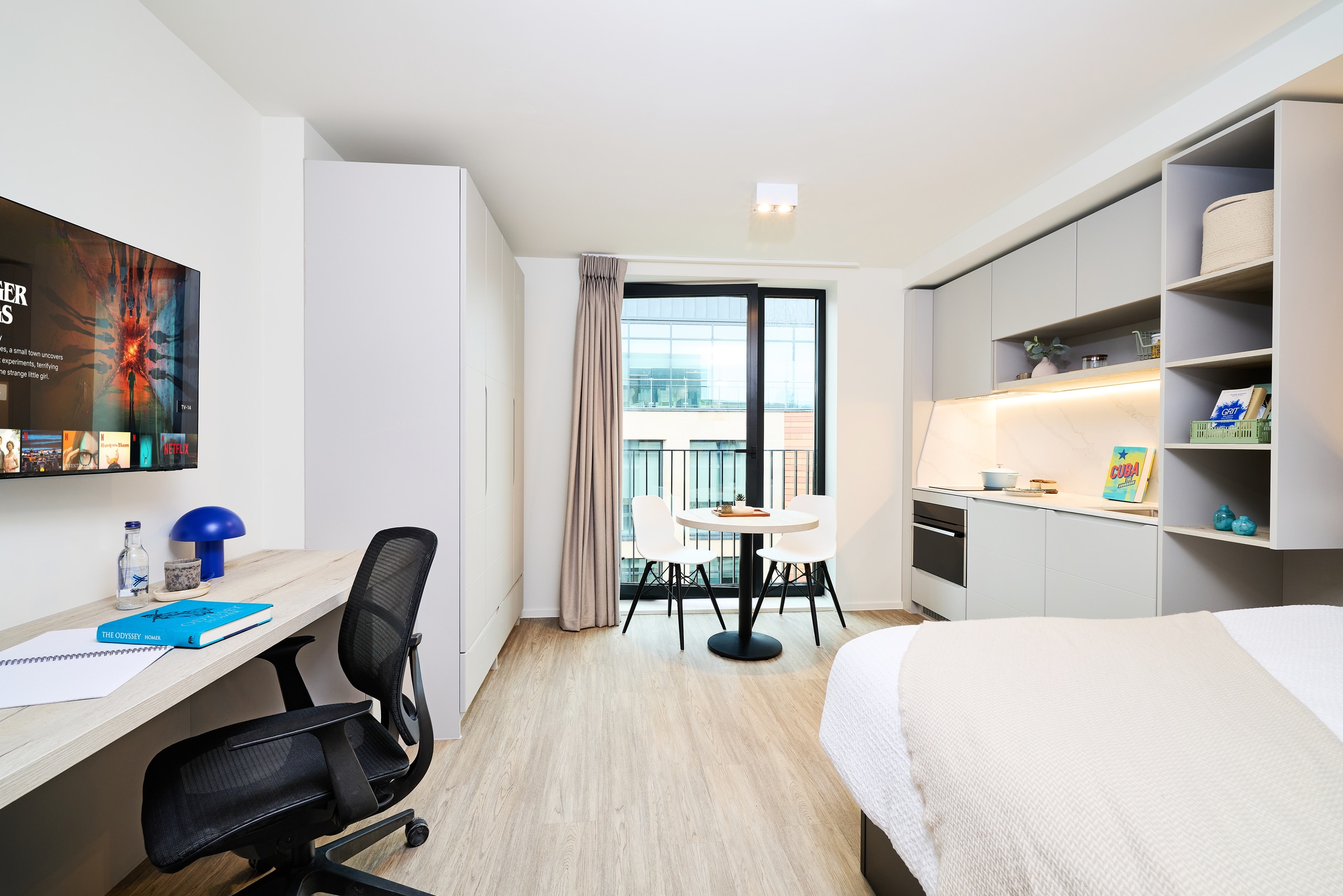 Modern studio apartment with a bed, desk, TV, kitchenette, and small dining table by a tall window. Light wood floors and minimalist decor create a bright, clean atmosphere.