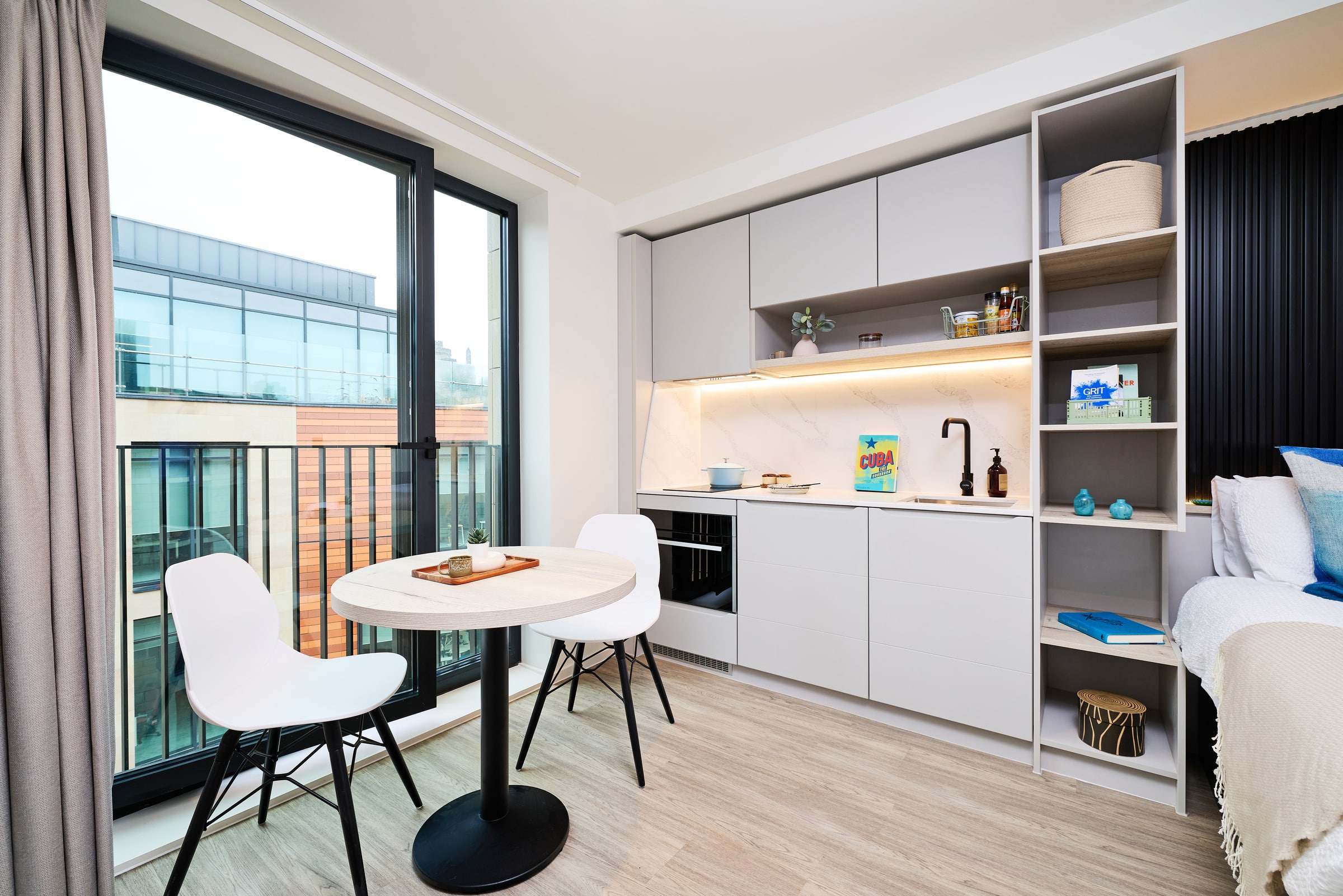 Modern studio apartment with a compact kitchen, round table with two white chairs, open shelving, and a large window overlooking a building. The space is bright and neatly organized.