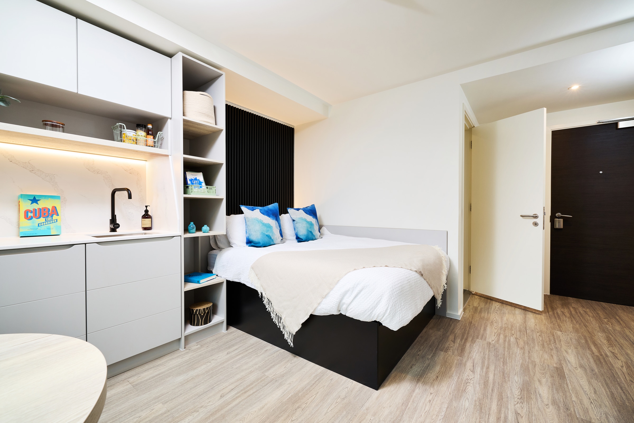 Modern studio apartment with a bed, white bedding, blue pillows, open shelving, a small kitchenette, and a light wood floor. The door is open, letting in light from the hallway.