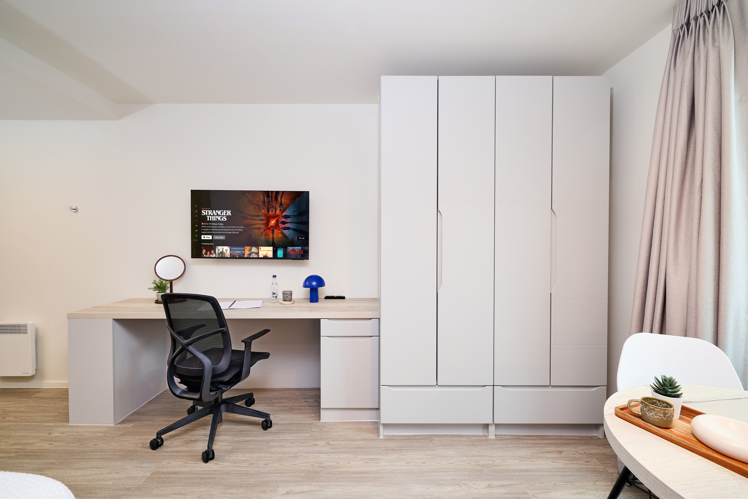 Modern workspace with a desk, office chair, TV on the wall, lamp, and mirror. Next to the desk is a large white wardrobe. Natural light enters through a window with gray curtains.