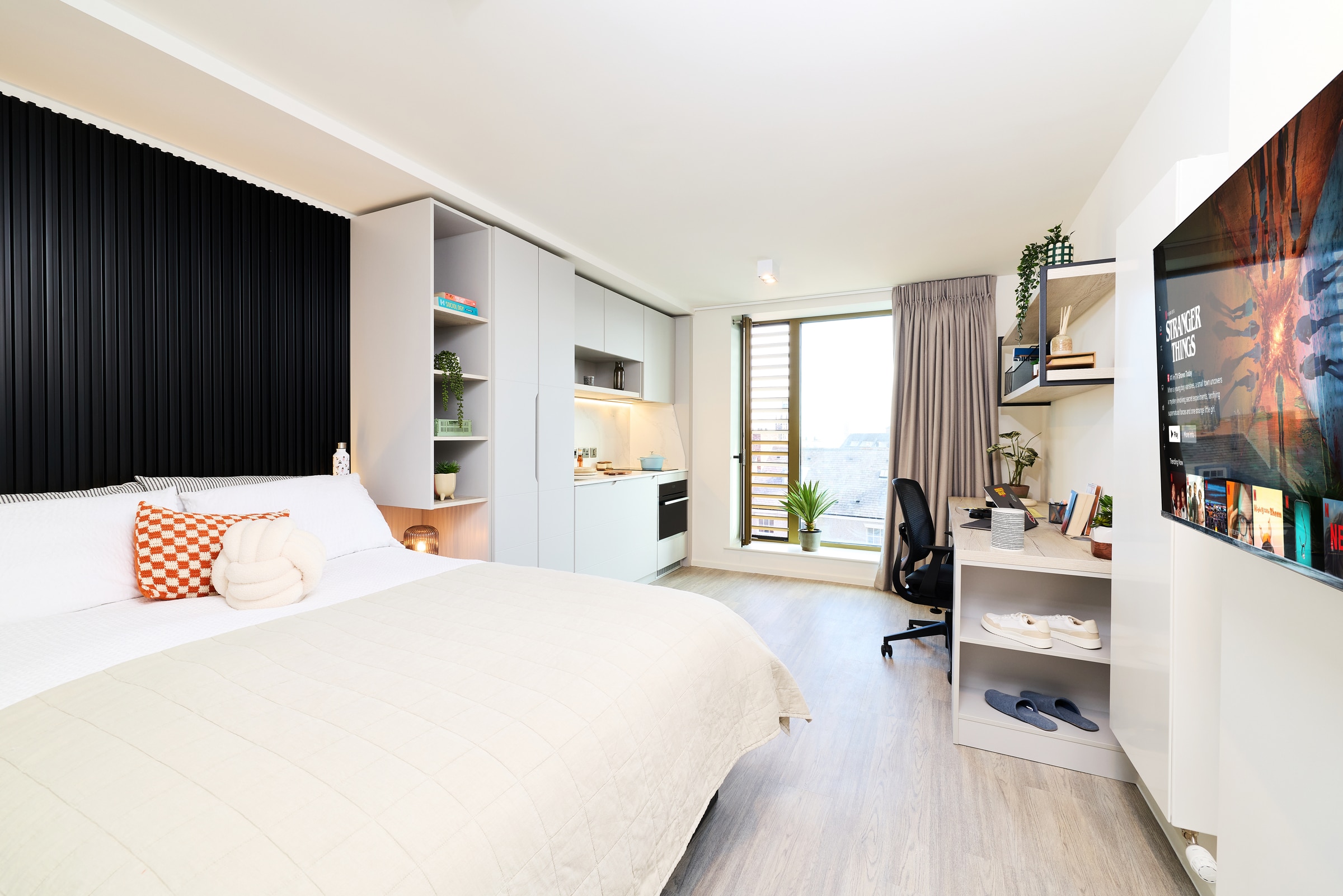 Modern bright bedroom with a large bed, desk, wall-mounted TV, open shelves, kitchenette, and a window with city views and a potted plant.