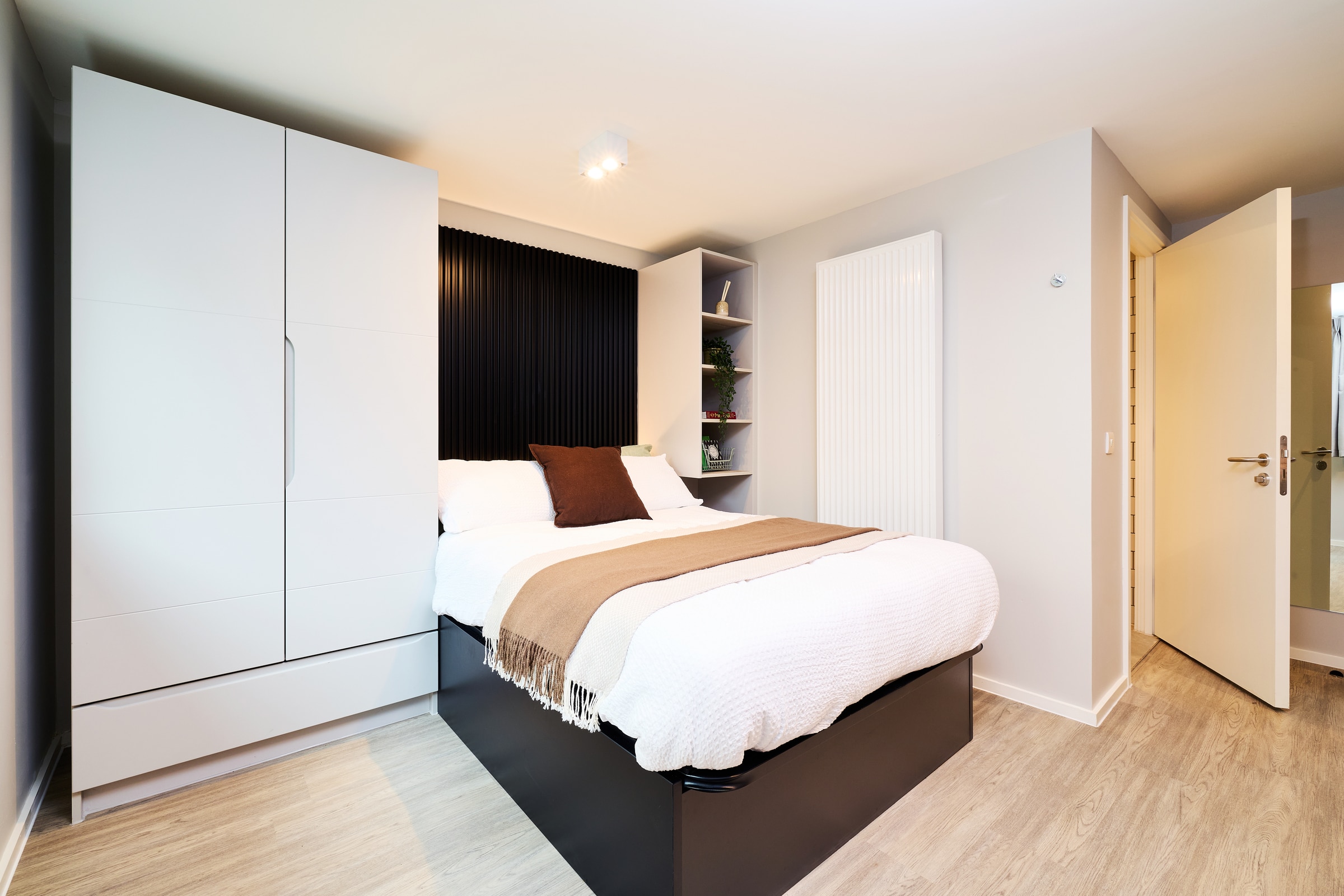 Modern bedroom with a neatly made bed, white bedding, beige throw, and brown cushion. There’s a white wardrobe, open shelves with decor, light wood flooring, and an open door leading to another room.