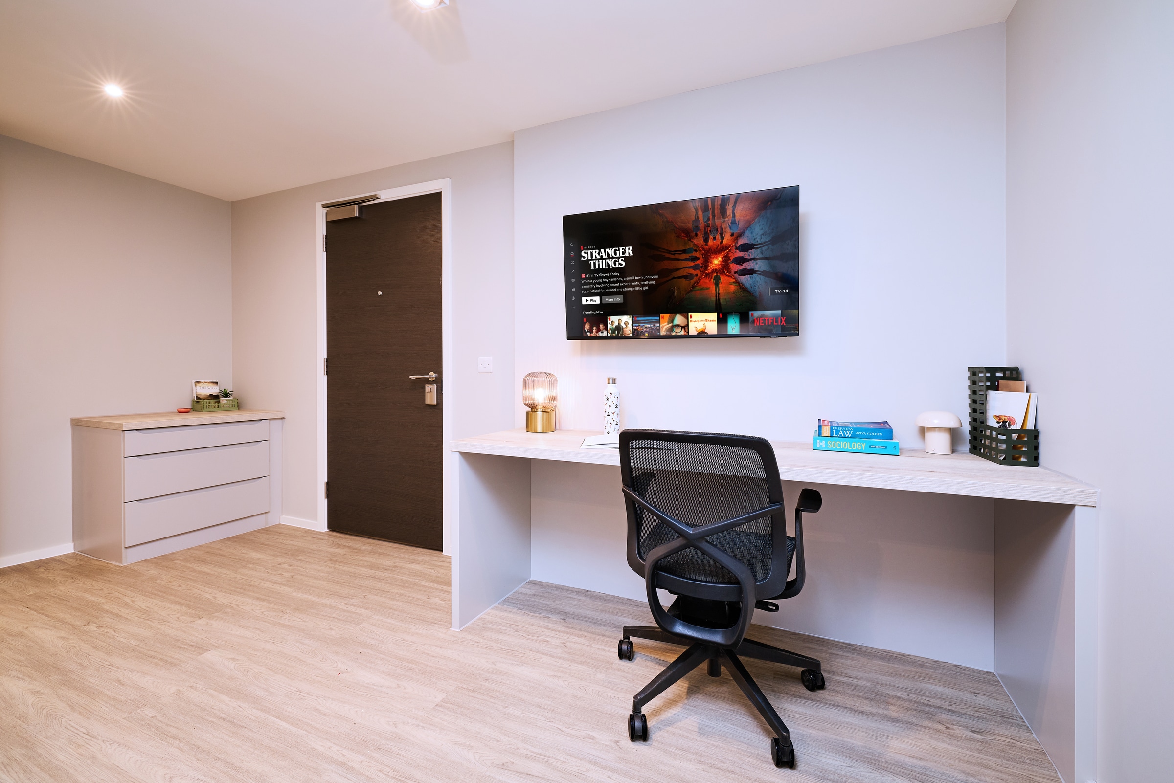 Modern home office with a black chair at a desk, books, lamp, and decor on the surface. A TV mounted on the wall displays a streaming menu. Wooden floors and white walls complete the clean look.