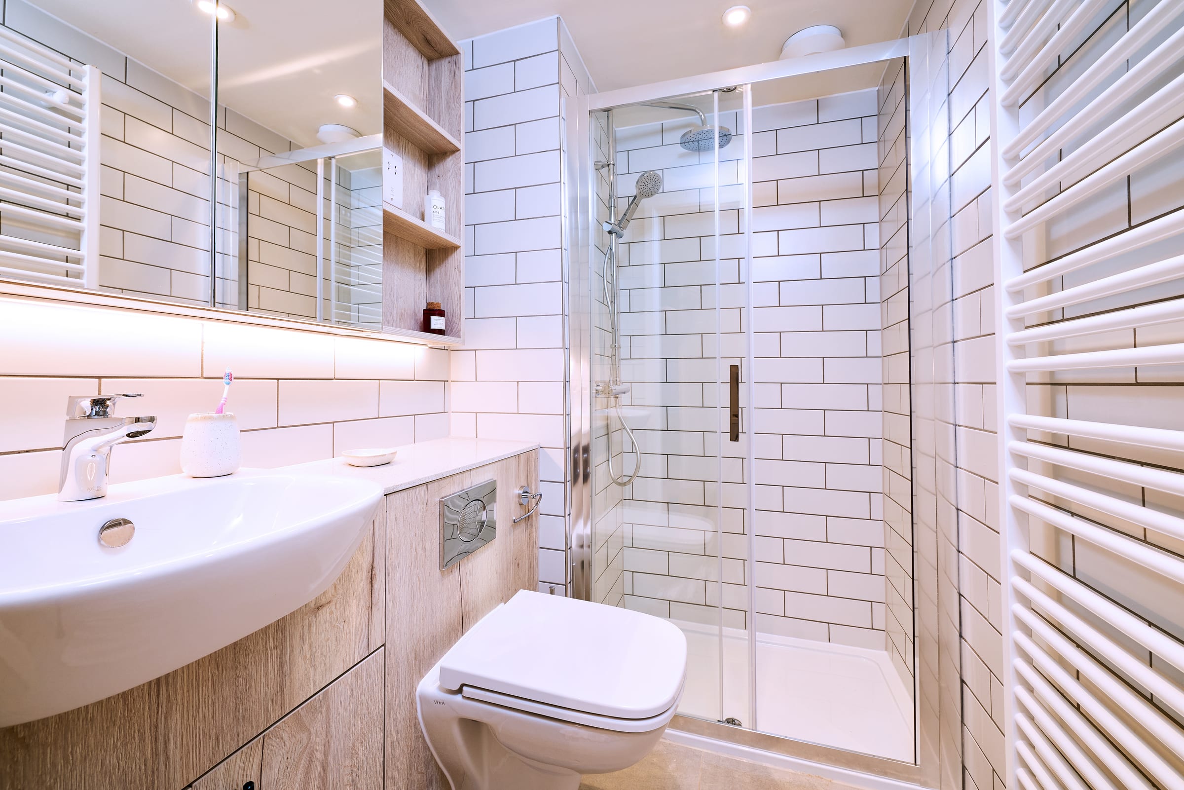 Modern bathroom with white subway tiles, glass shower enclosure, wall-mounted toilet, sink with a mirror above, open wooden shelves, and a white towel warmer on the right wall.