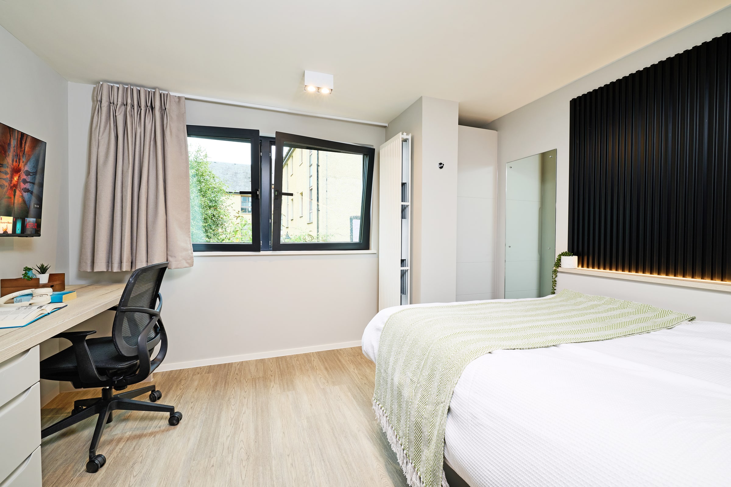 Modern bedroom with a bed, green blanket, desk with chair, TV, window with beige curtain, wardrobe, and vertical black wall panels. The room has wooden flooring and a clean, minimalist design.