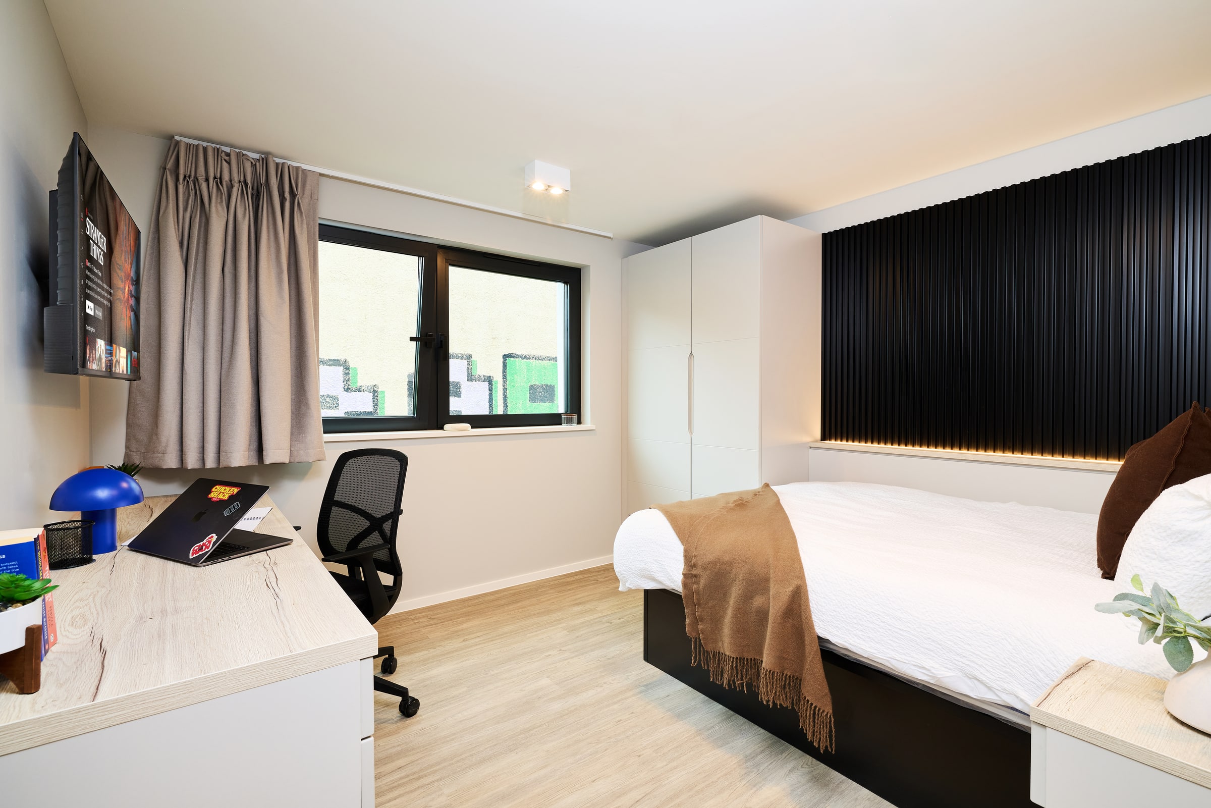 Modern bedroom with a bed, brown blanket, desk with laptop, desk chair, lamp, wardrobe, window with gray curtains, and black wall paneling behind the bed.