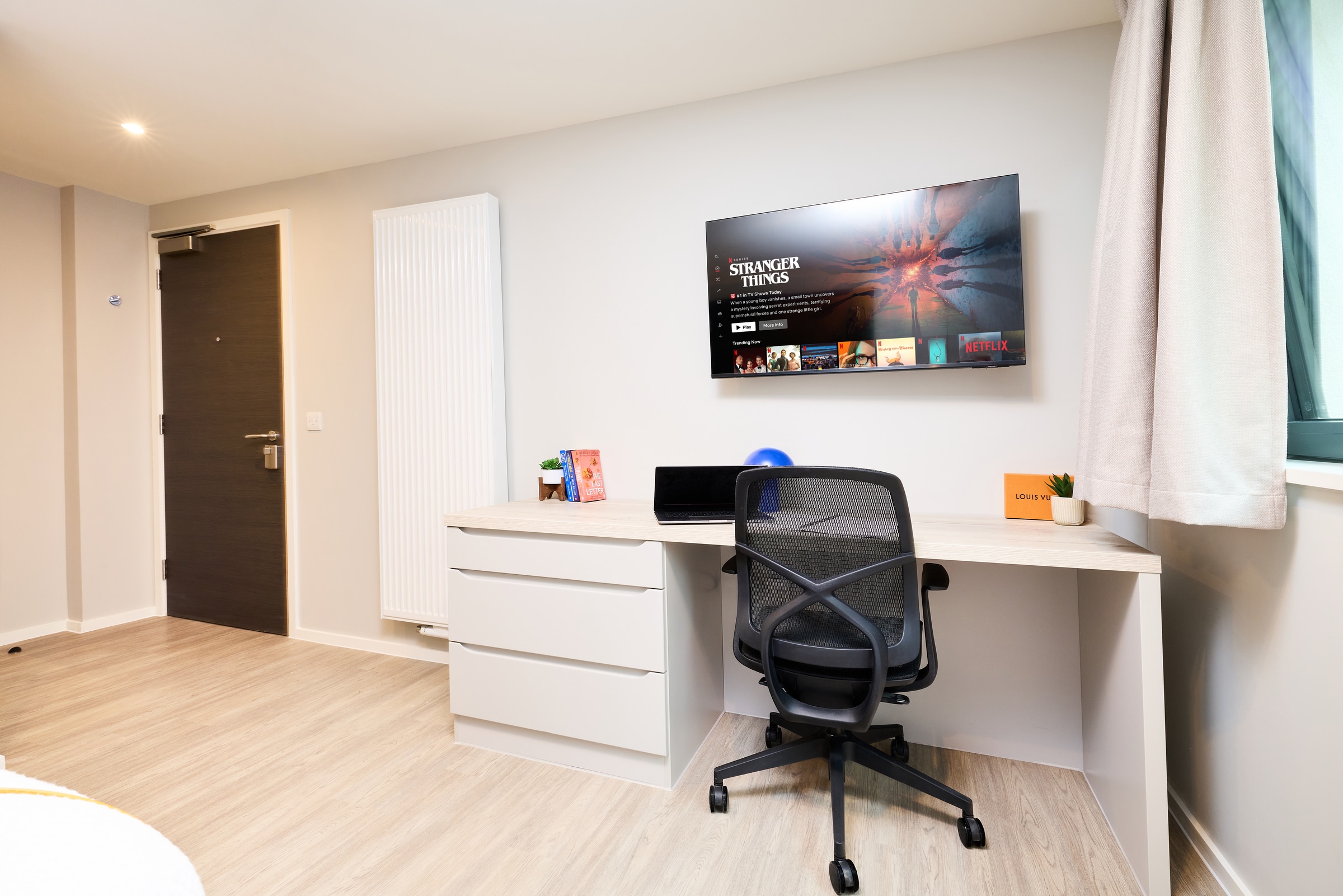 A modern study space with a desk, office chair, laptop, and decor. A TV on the wall displays “Stranger Things.” A window with curtains lets in natural light. There’s a closed door and a radiator on the left.