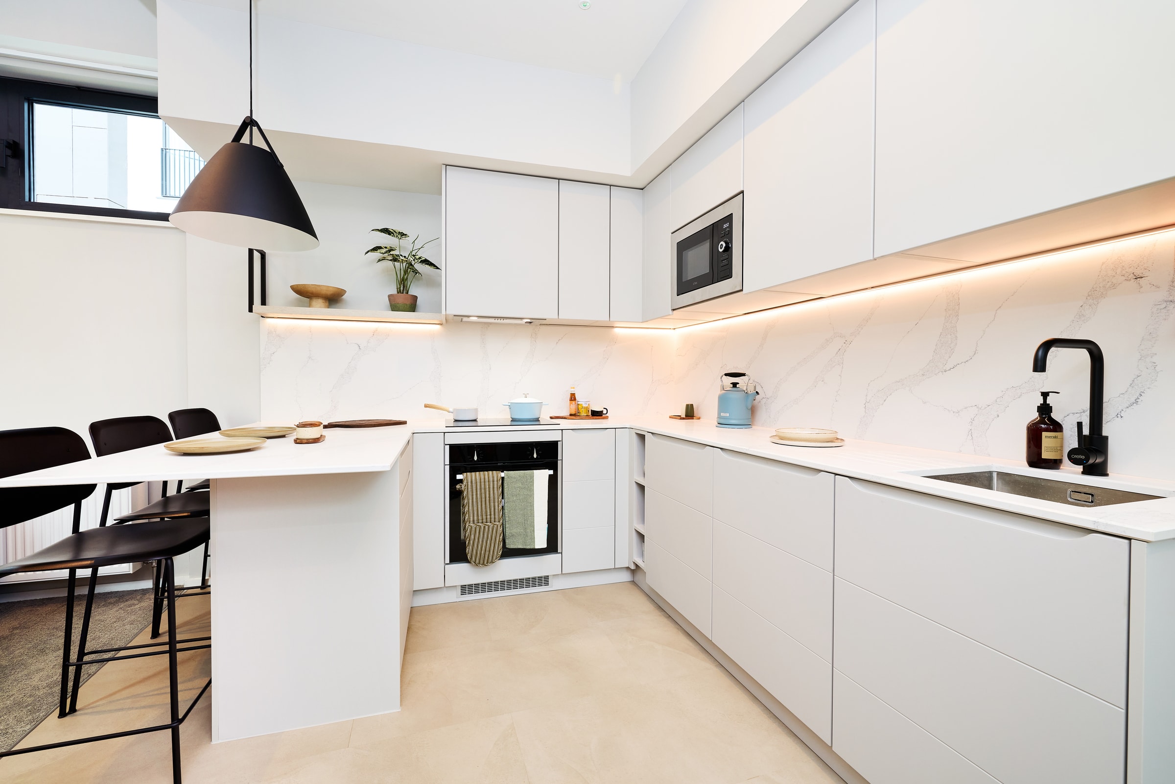 Modern white kitchen with under-cabinet lighting, marble backsplash, built-in oven, microwave, sink, and a breakfast bar with three black stools. Minimalist decor includes dishes, plants, and a pendant light.