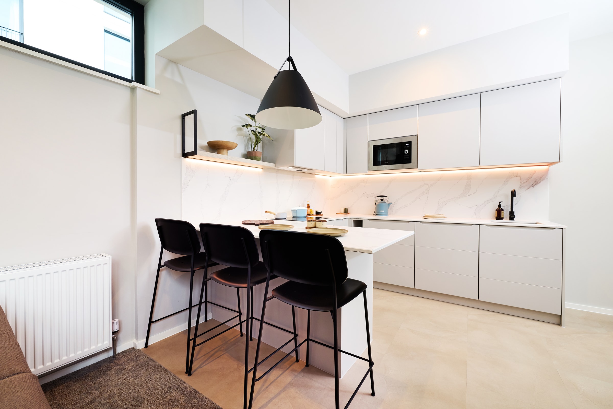 Modern white kitchen with minimalist cabinets, an island with three black barstools, pendant light, built-in microwave, and subtle decor including plants and tableware. Warm under-cabinet lighting brightens the space.