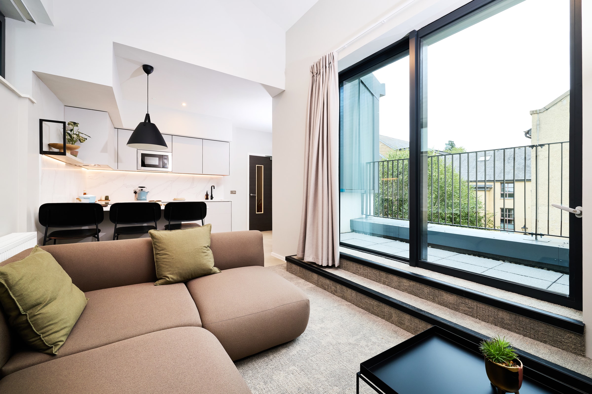 Modern living room with a brown sofa, green pillows, and a small black table. Open kitchen with bar stools is in the background. Large glass doors open to a balcony with a view of trees and buildings.