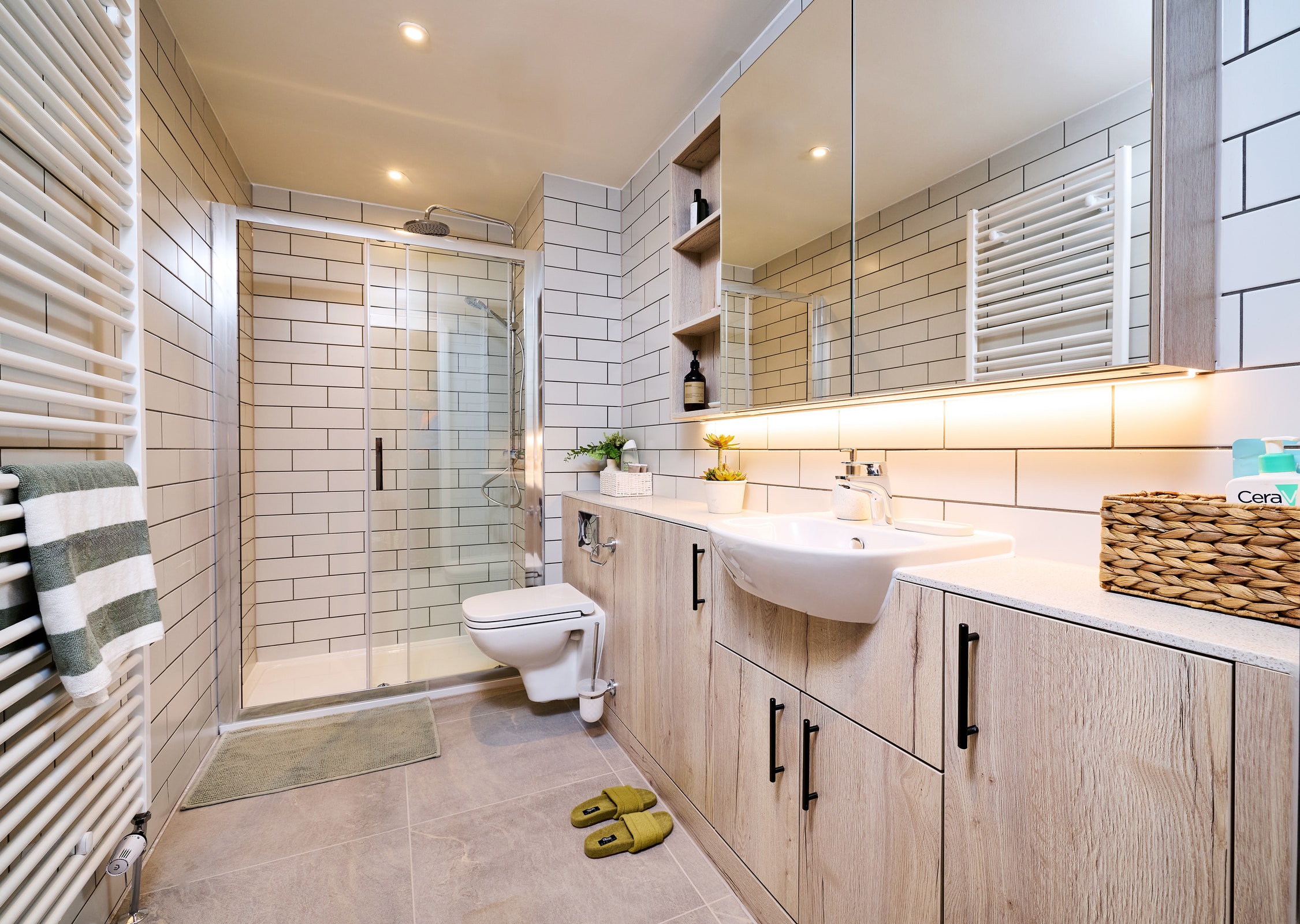 Modern bathroom with light wood cabinets, wall-mounted toilet, glass shower, white sink, subway tile walls, woven basket, plants, and slippers on the floor. Large mirror with shelves and bright lighting.