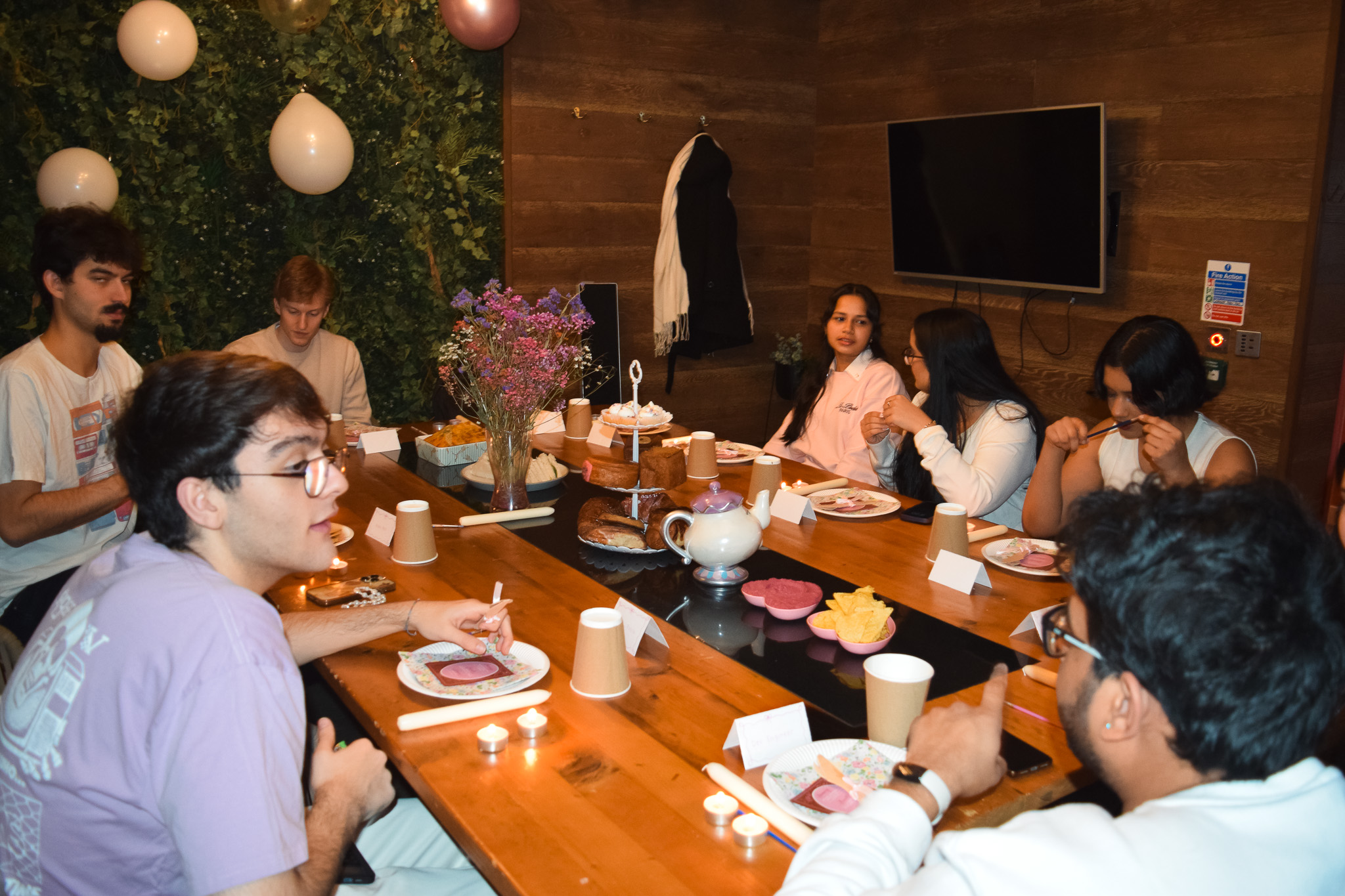 Seven people sit around a wooden table with food and drinks, chatting and eating. The room has balloons, greenery on the wall, and a TV. Place cards and candles decorate the table as laughter fills the air and sparkly skirts catch the light, adding to the festive atmosphere.