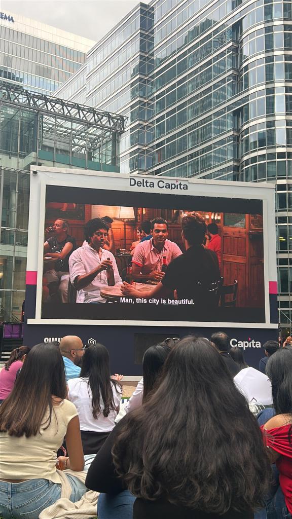 People sit outdoors watching a movie on a large screen in front of modern glass buildings. The screen displays a scene with three people sitting at a table and English subtitles reading, “Man, this city is beautiful.” As part of the Vita Student Student Stories series, this open-air cinema event brings together students to celebrate city life and shared experiences.