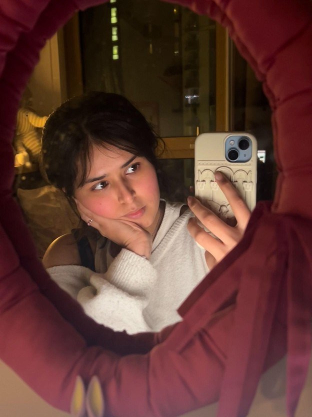 A person wearing a white sweater takes a mirror selfie, resting their chin on their hand and looking thoughtfully to the side—capturing one of those quiet student stories that speak of growing pains. The mirror is framed with a padded, deep red cover and ribbon.