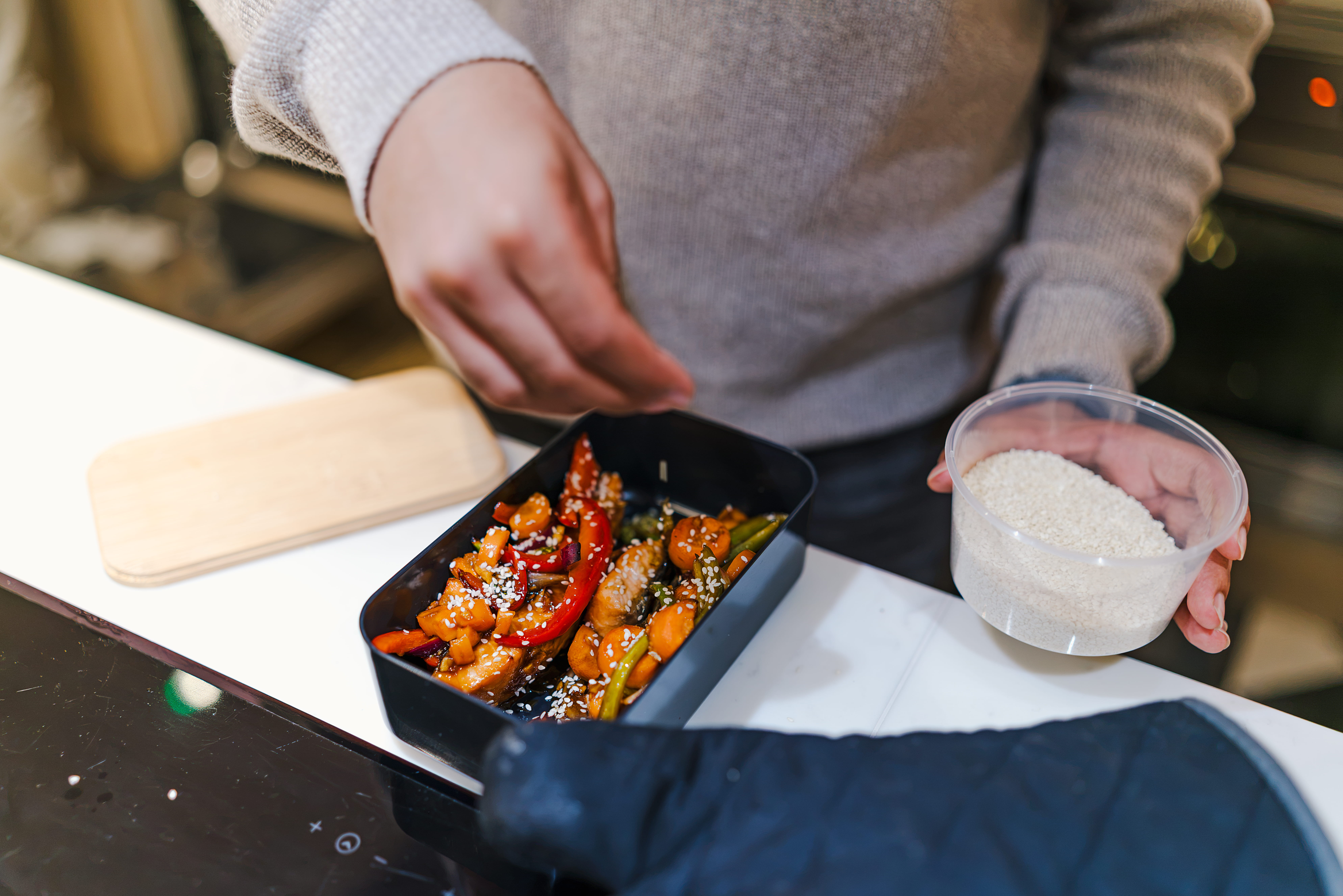 A person sprinkles sesame seeds onto a meal in a black container, adding the seeds from a small clear bowl. The dish appears to contain vegetables and meat, garnished with red chili peppers.