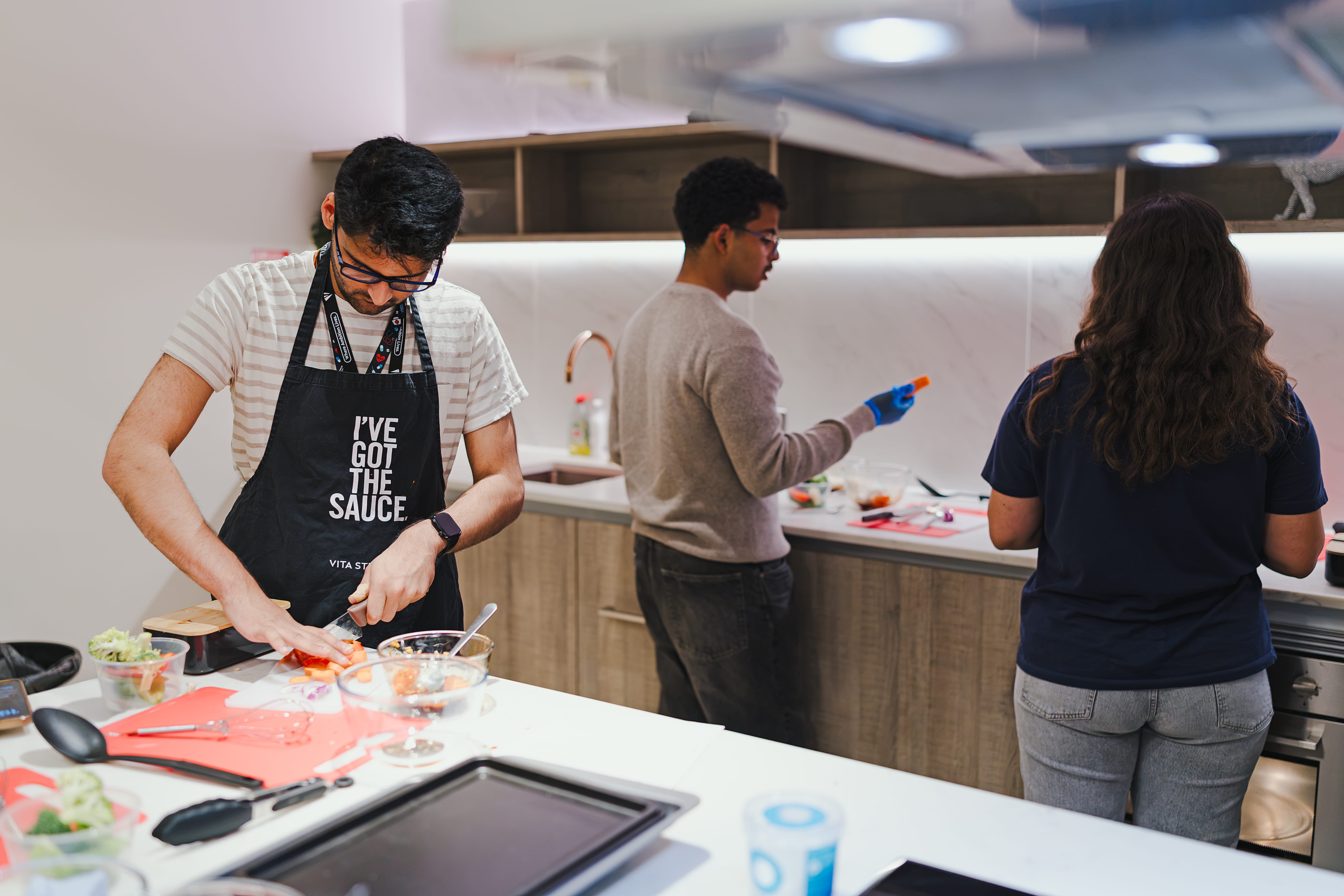 Three people prepare food in a modern kitchen. One man wears an apron that says “I’ve got the sauce” while cutting ingredients. Two others work at the counter, focused on their own cooking tasks.