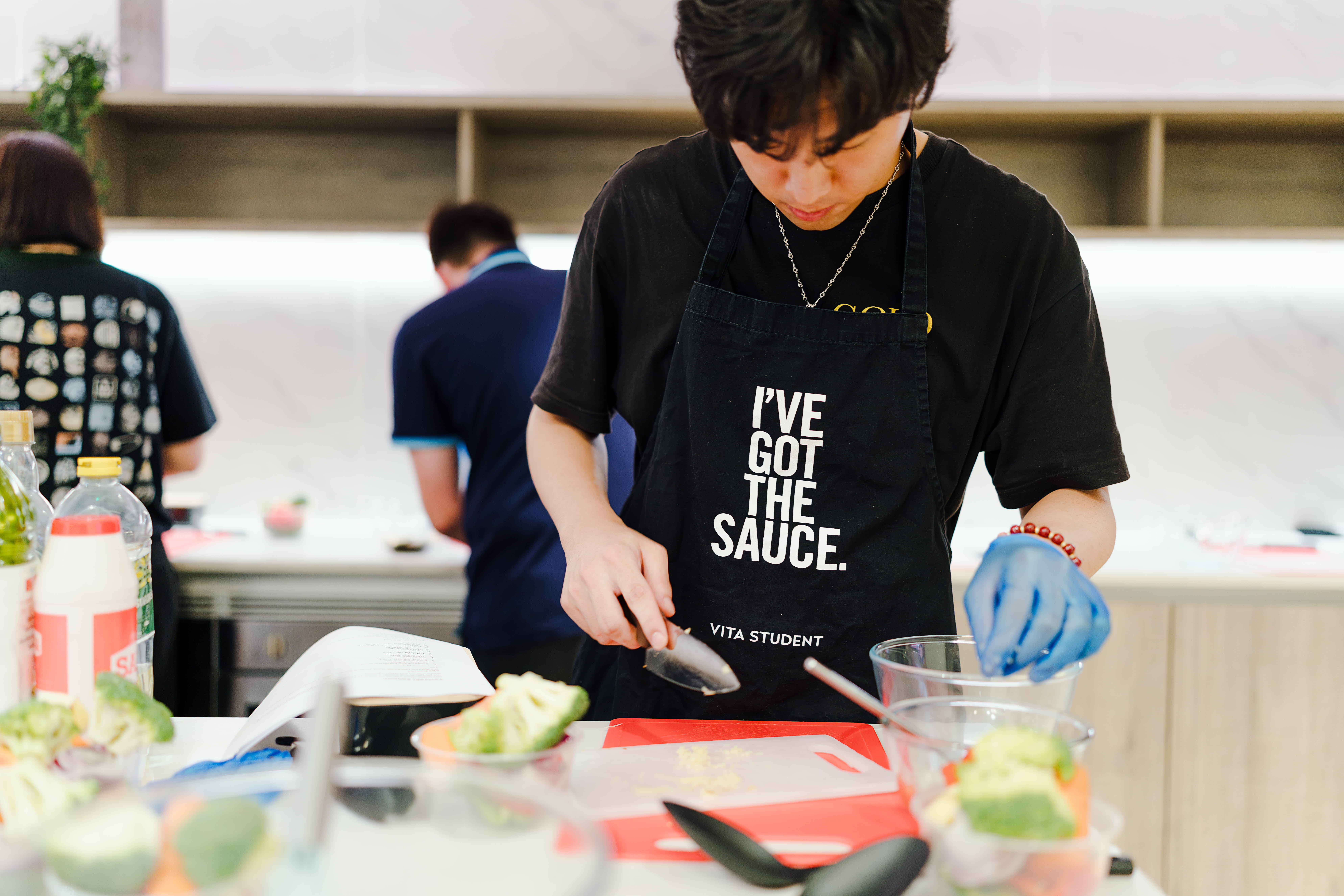 A person wearing a black apron that says “I’VE GOT THE SAUCE.” chops ingredients in a kitchen, focused on preparing food. Various vegetables and utensils are on the counter.