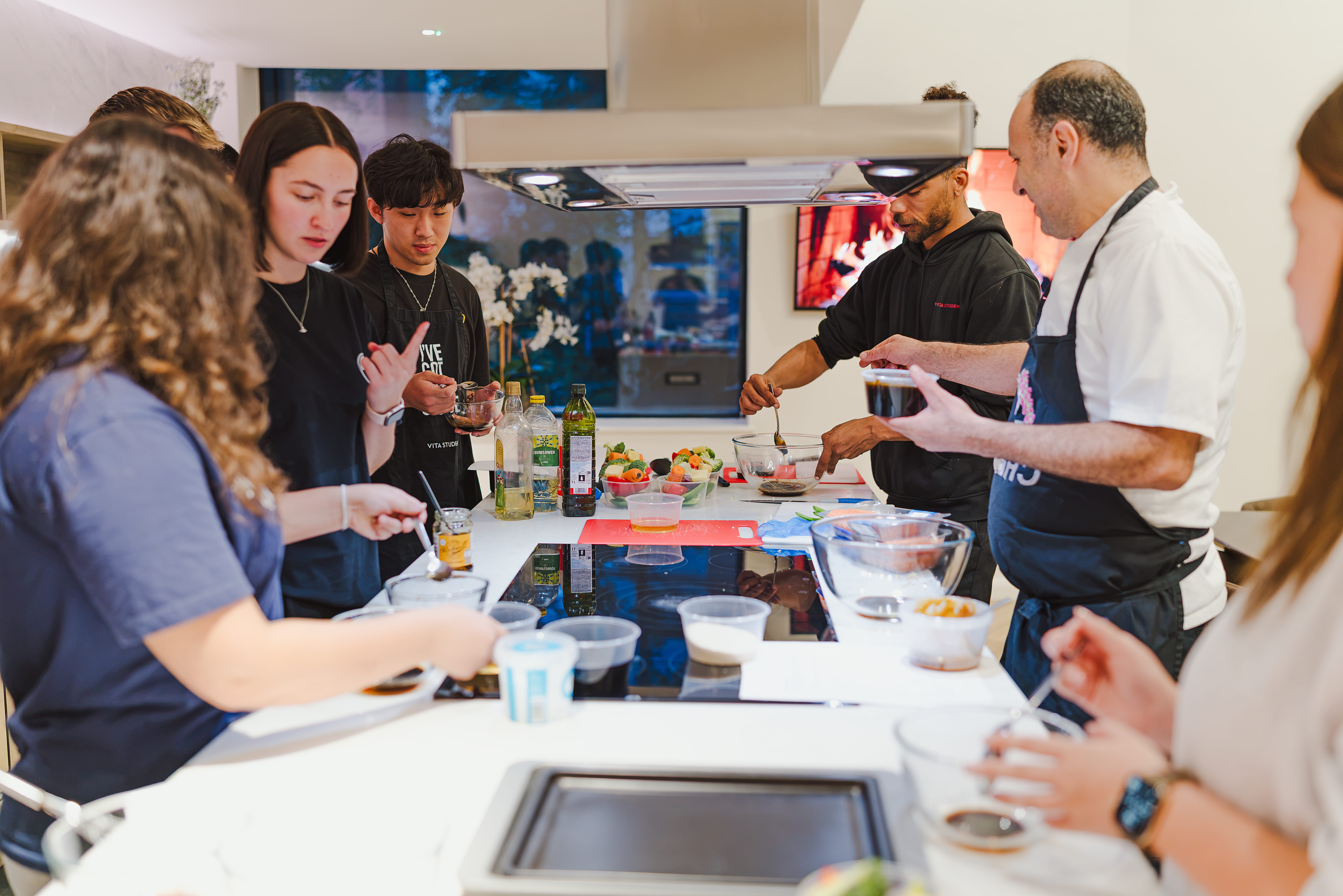 Six people gather around a kitchen island, preparing food together. Various ingredients, bowls, and utensils are spread out as a chef demonstrates a technique. The atmosphere appears lively and interactive.