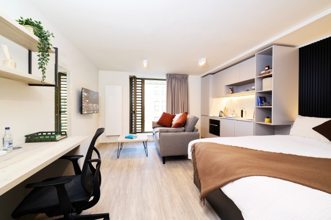 A modern studio apartment with a bed, sofa, desk, wall shelves, TV, and kitchen area. Neutral colors, wooden floor, large window with curtains, and contemporary lighting create a clean and cozy atmosphere.