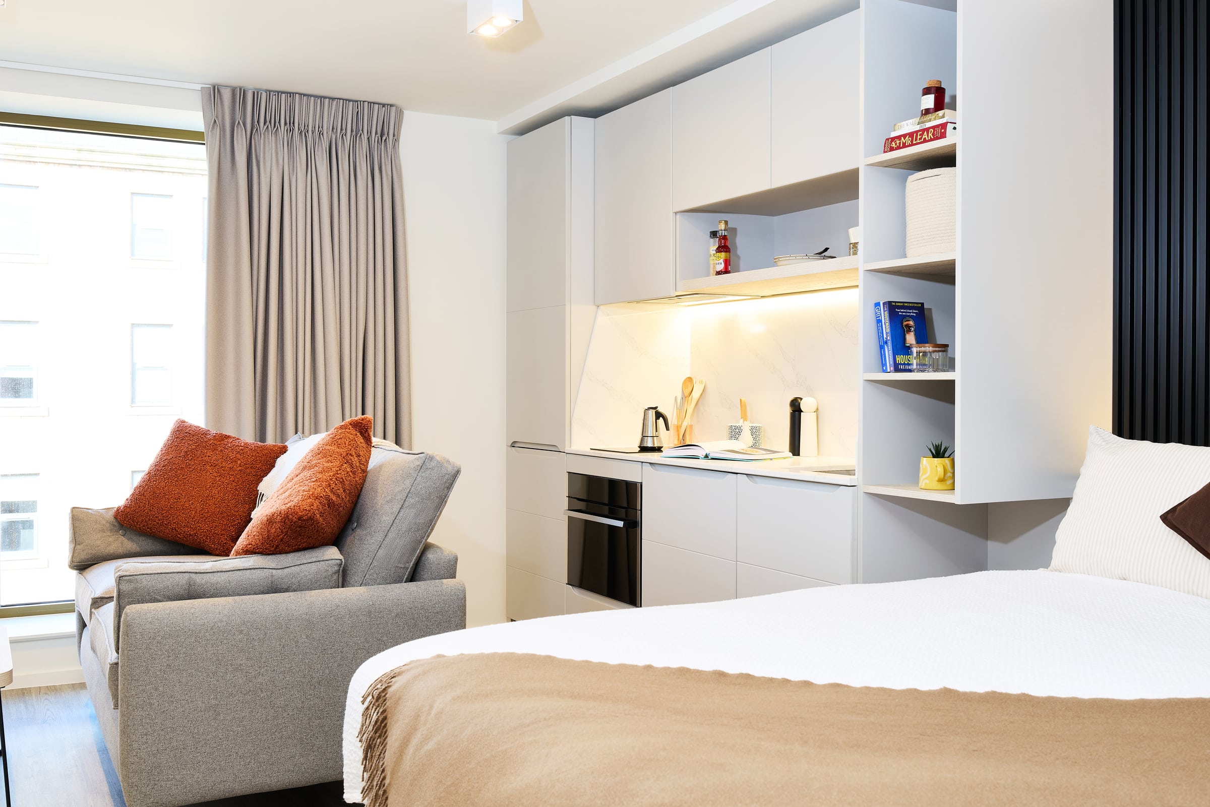 Modern studio apartment with a bed, gray sofa with orange pillows, compact kitchen, open shelves, and large window with curtains letting in natural light.