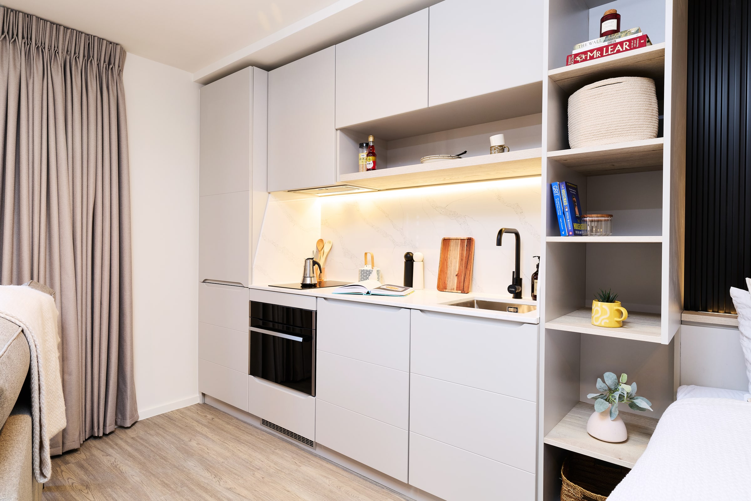 Modern kitchenette with light gray cabinets, open shelves, a built-in oven, sink, stovetop, cutting boards, and various kitchen items. Wooden floor and beige curtains complete the cozy, minimalist space.