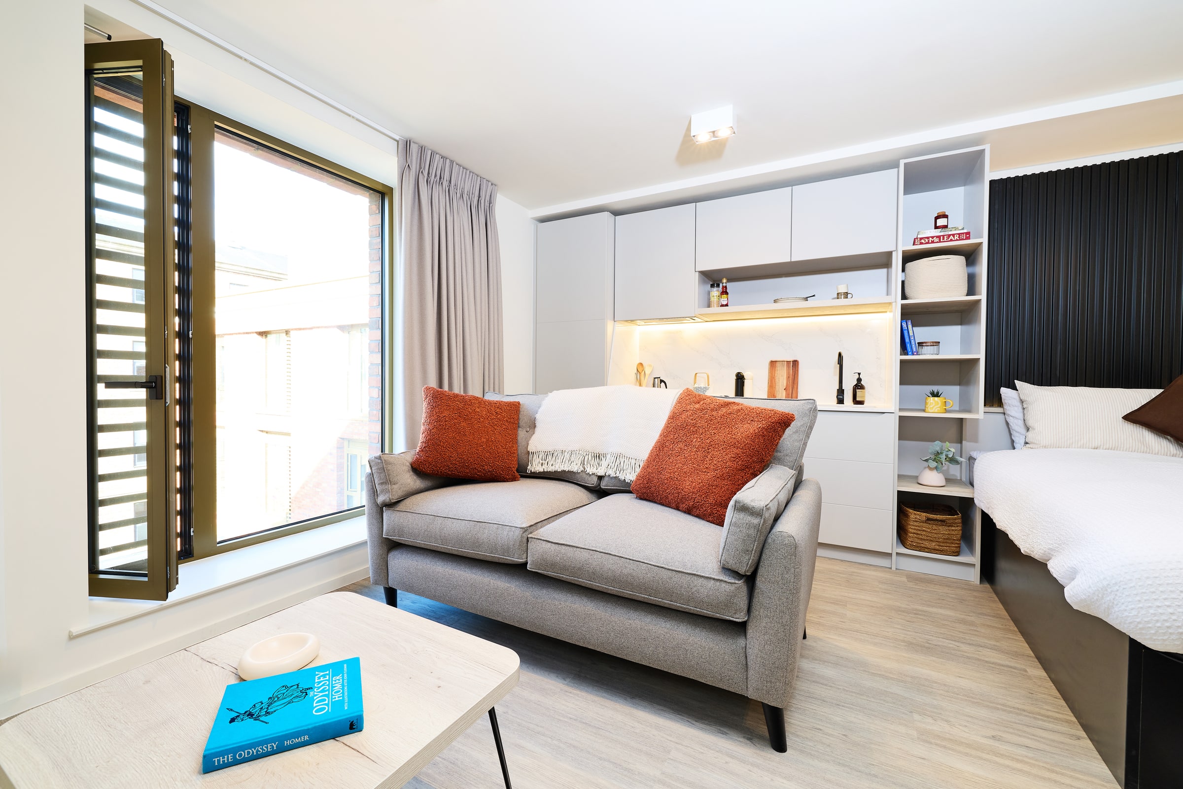 A modern studio with a gray sofa, orange cushions, and a blanket. There’s a bed, shelves, a window with blinds, and a table with a book and a dish. The space is bright with light wood floors and minimalist decor.