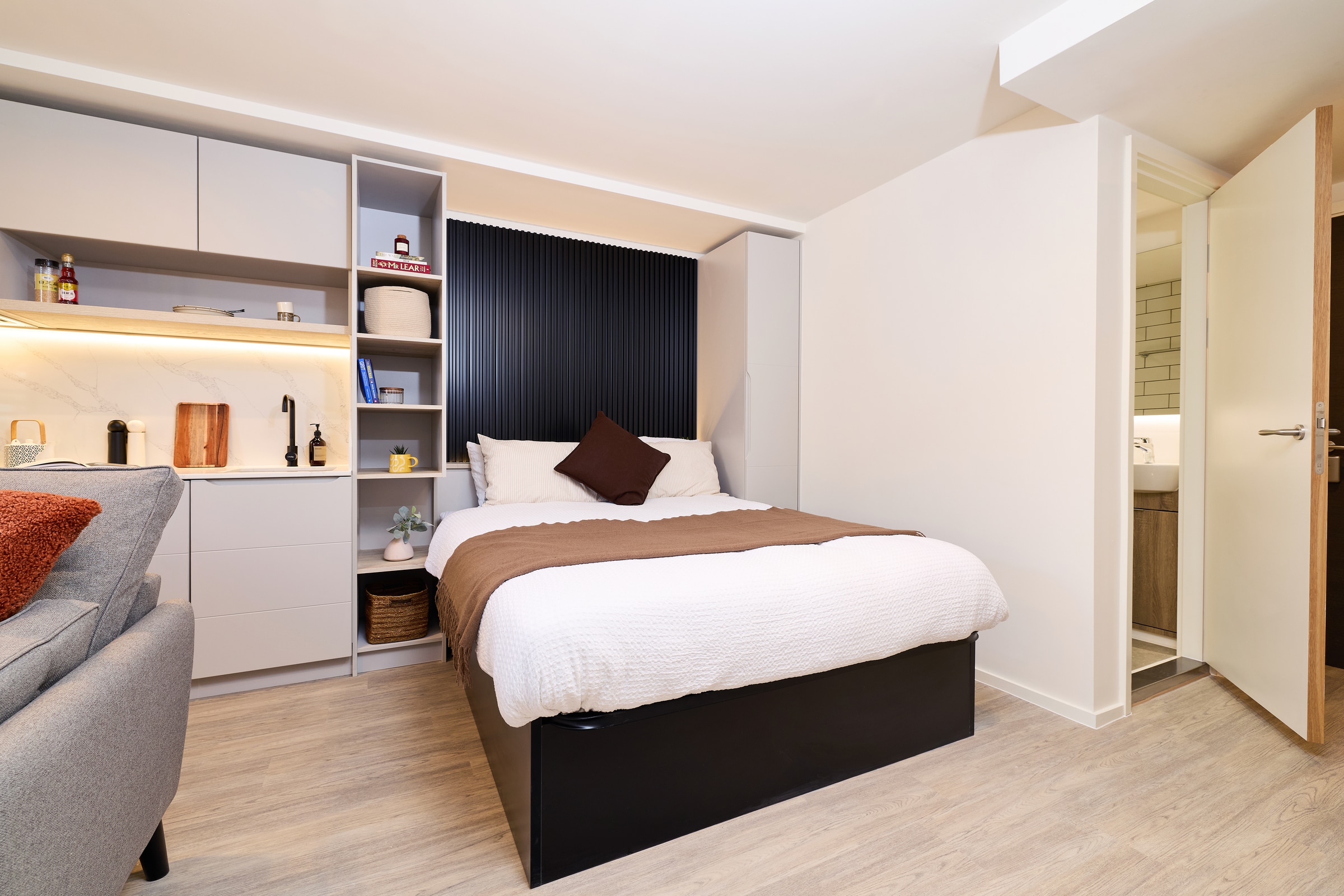 A modern studio bedroom with a double bed, open shelving, a partial grey sofa, and a view into a bathroom with tiled walls, all in neutral tones and tidy arrangement.
