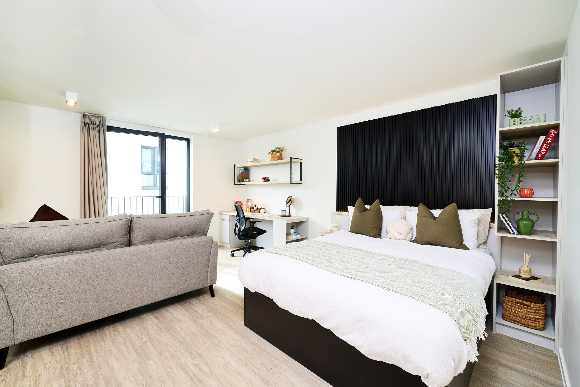Modern studio apartment with a bed, pillows, shelves, a desk with a chair, a sofa, large window, and light wood flooring. The space is bright and neatly organized.