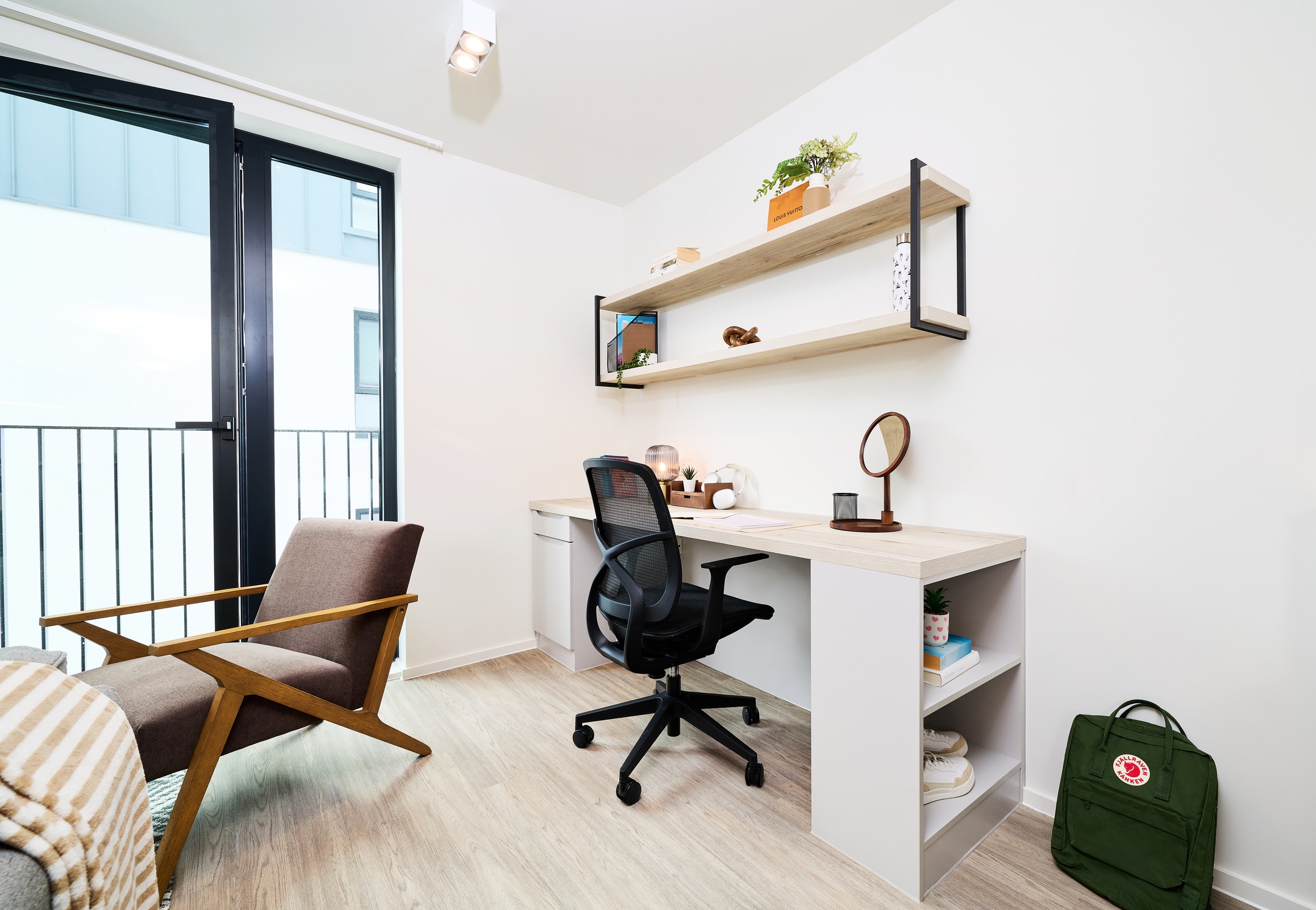 Modern, minimalist home office with a wooden desk, black chair, wall shelves with plants, and a lamp. A green backpack sits on the floor, and a large window lets in natural light.