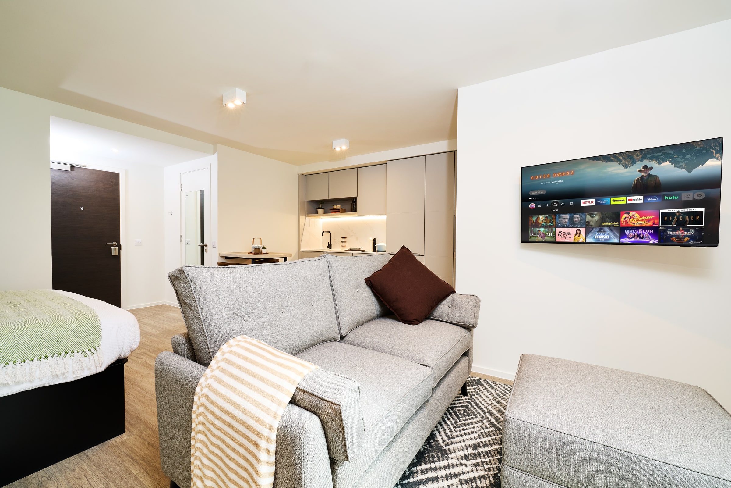A modern studio apartment with a gray sofa, TV mounted on the wall, small kitchen area, bed partially visible, light wood flooring, and neutral decor.