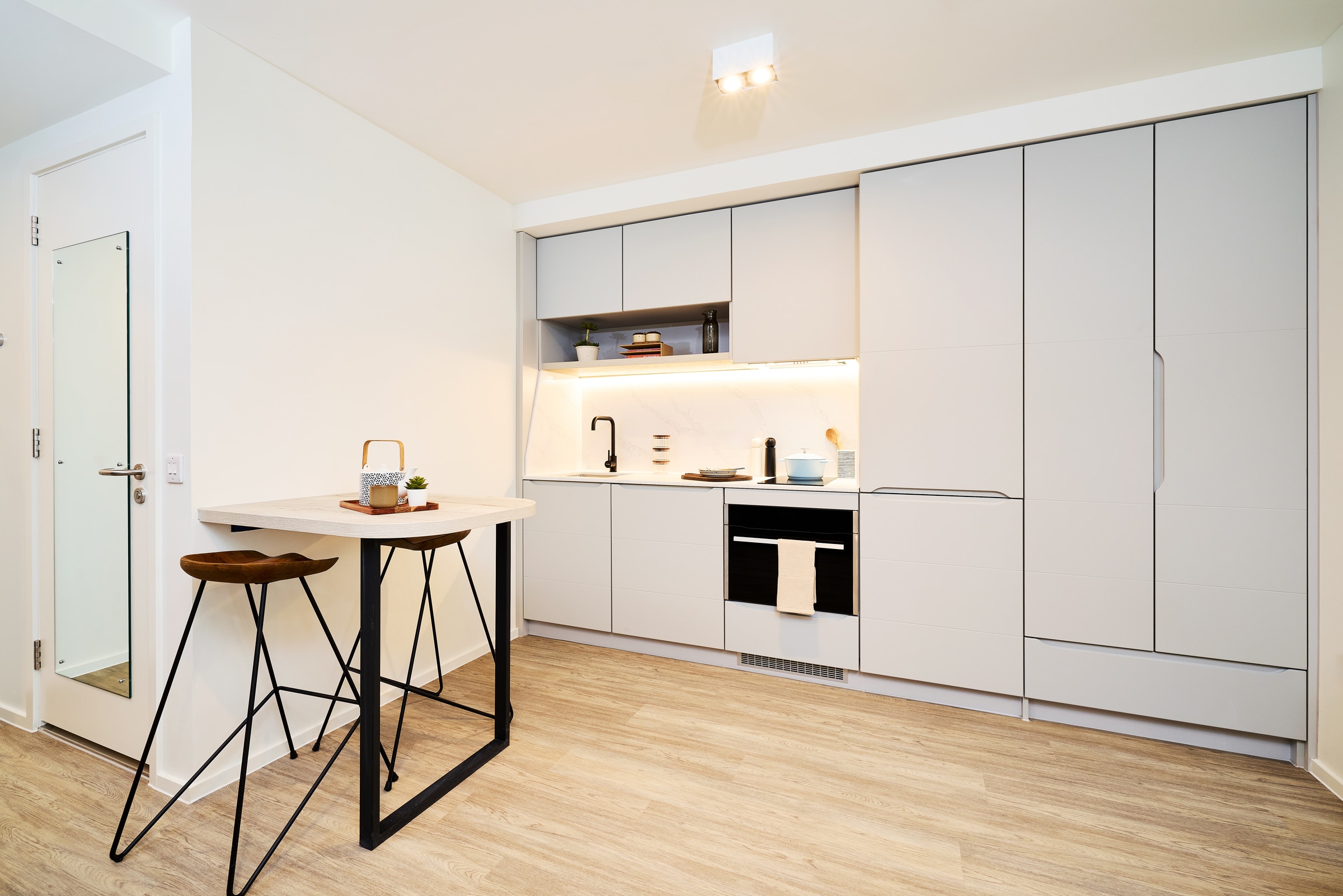 Modern kitchen with light gray cabinets, built-in appliances, wooden floor, a small dining table with two stools, and minimal decor. The room is bright with a clean, minimalist design.