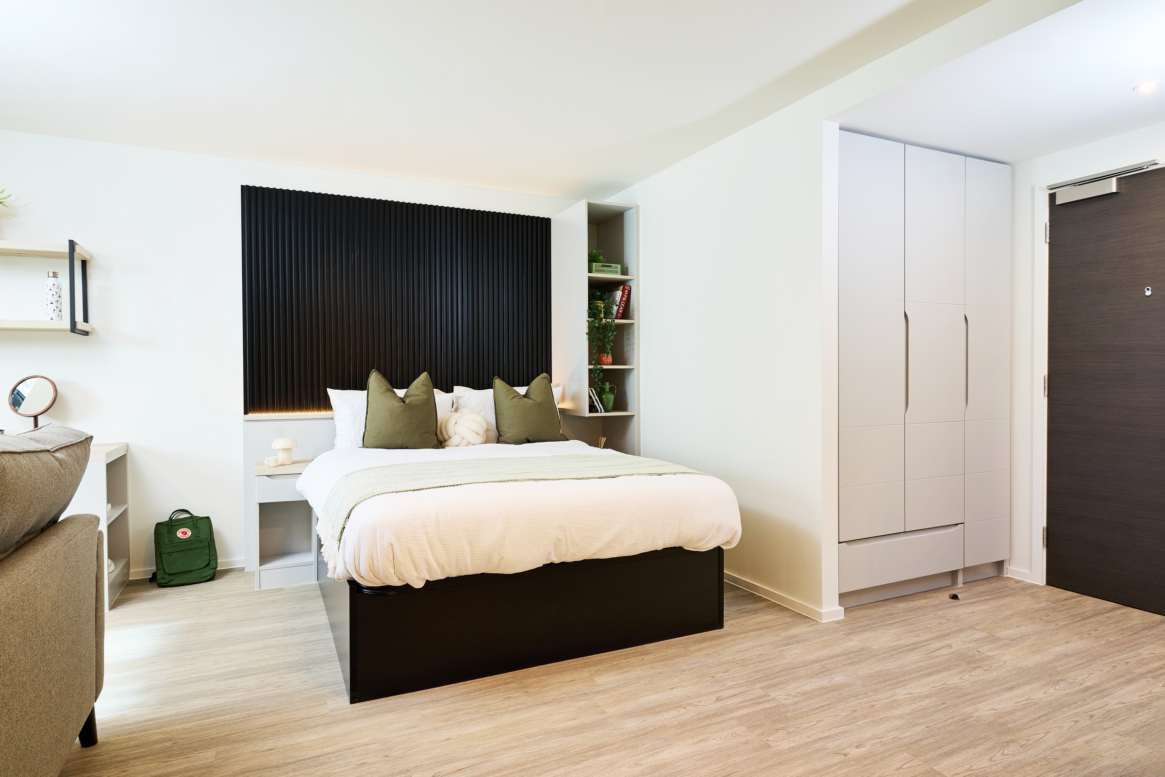 A modern, tidy bedroom with a double bed, green pillows, open shelves, a desk, built-in wardrobe, and light wood flooring. A green backpack is next to the bed.