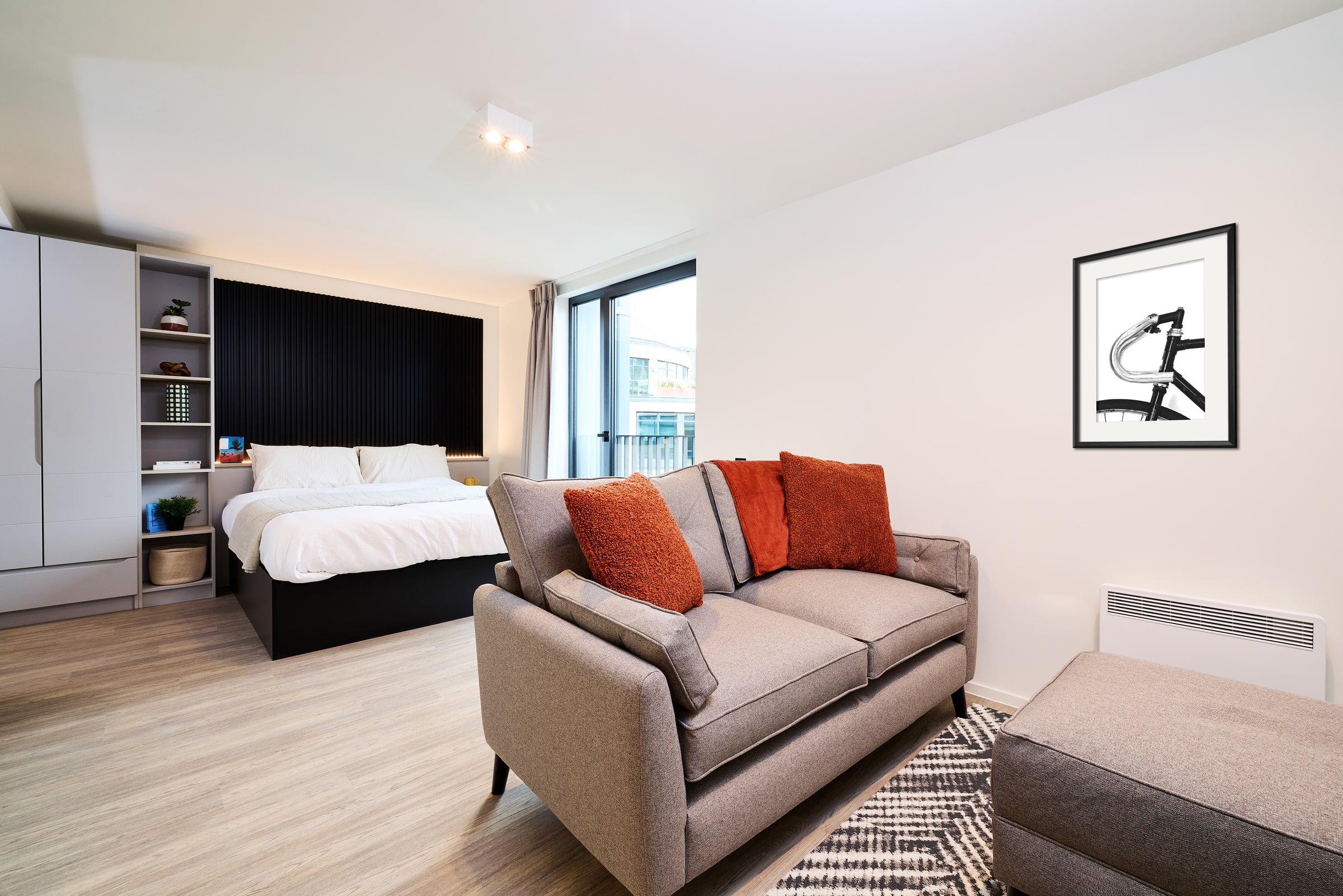 Modern studio apartment with a gray sofa, orange cushions, ottoman, large bed, framed artwork on the wall, light wood floors, and a window letting in natural light.