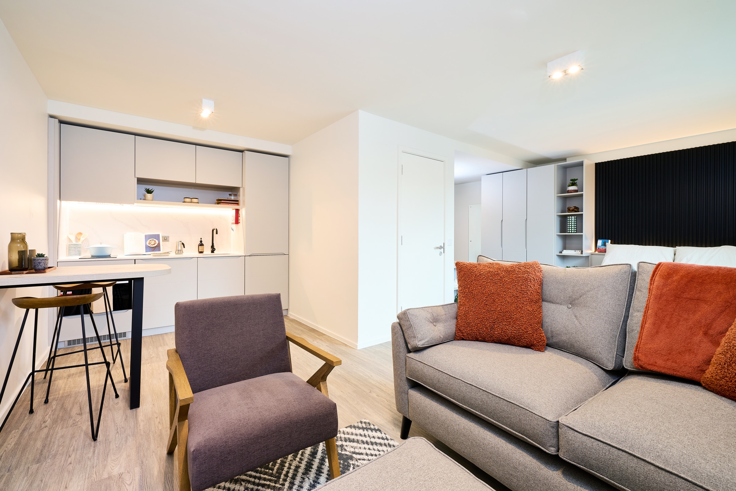 Modern open-plan studio apartment with a gray sofa, orange pillows, armchair, small kitchen area, bar table, and bright lighting. Minimalist decor and light wood flooring.