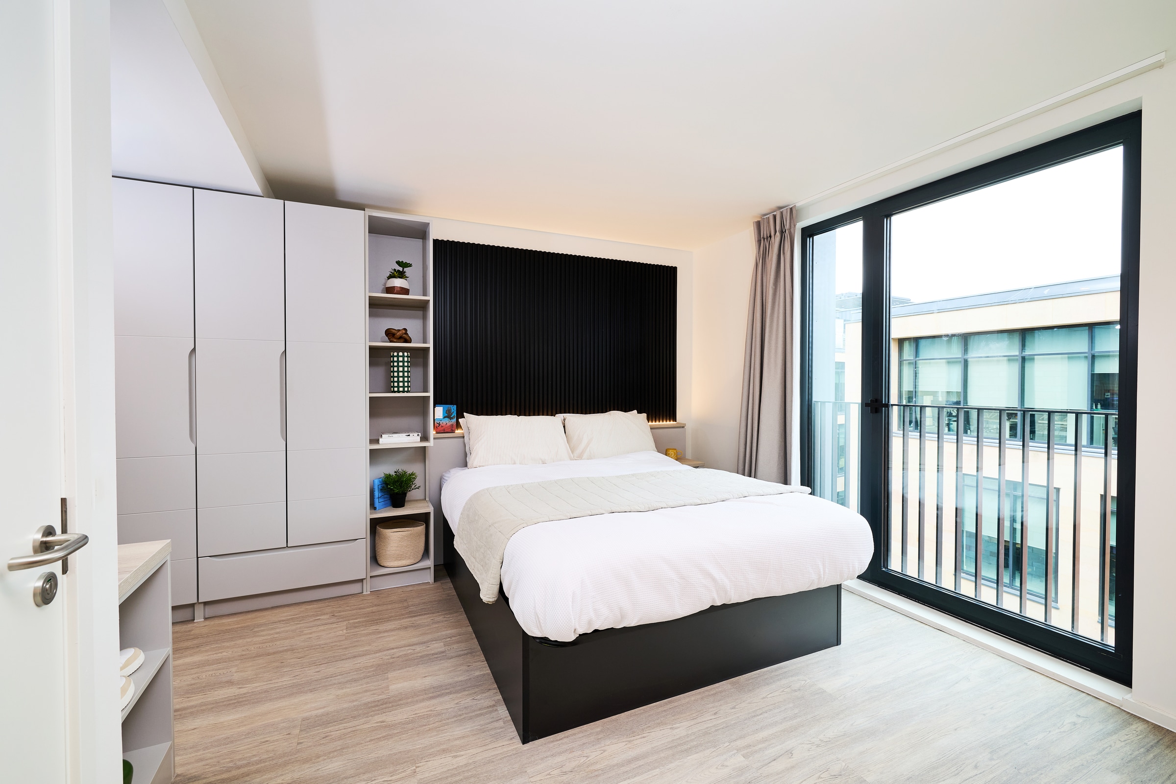 Modern bedroom with a large bed, white bedding, built-in closets, shelves with decor, a black accent wall, wood flooring, and a floor-to-ceiling window with a view of another building and a small balcony.
