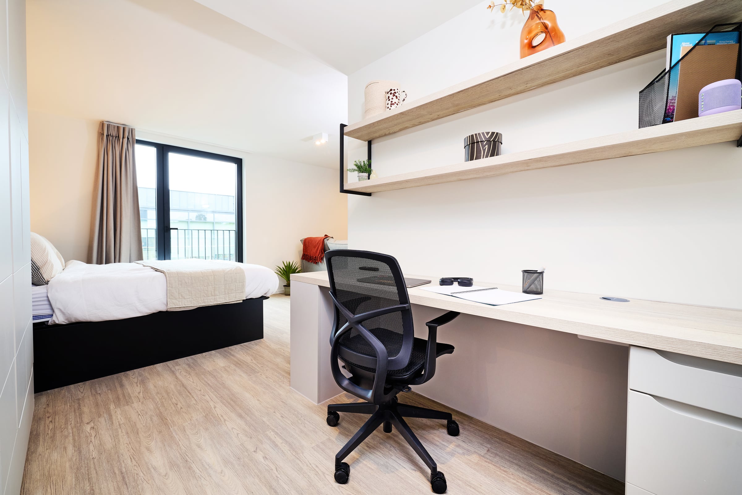 A modern, minimalist bedroom features a neatly made bed, large window, desk with a black chair, shelves with decor items, and light wood flooring.