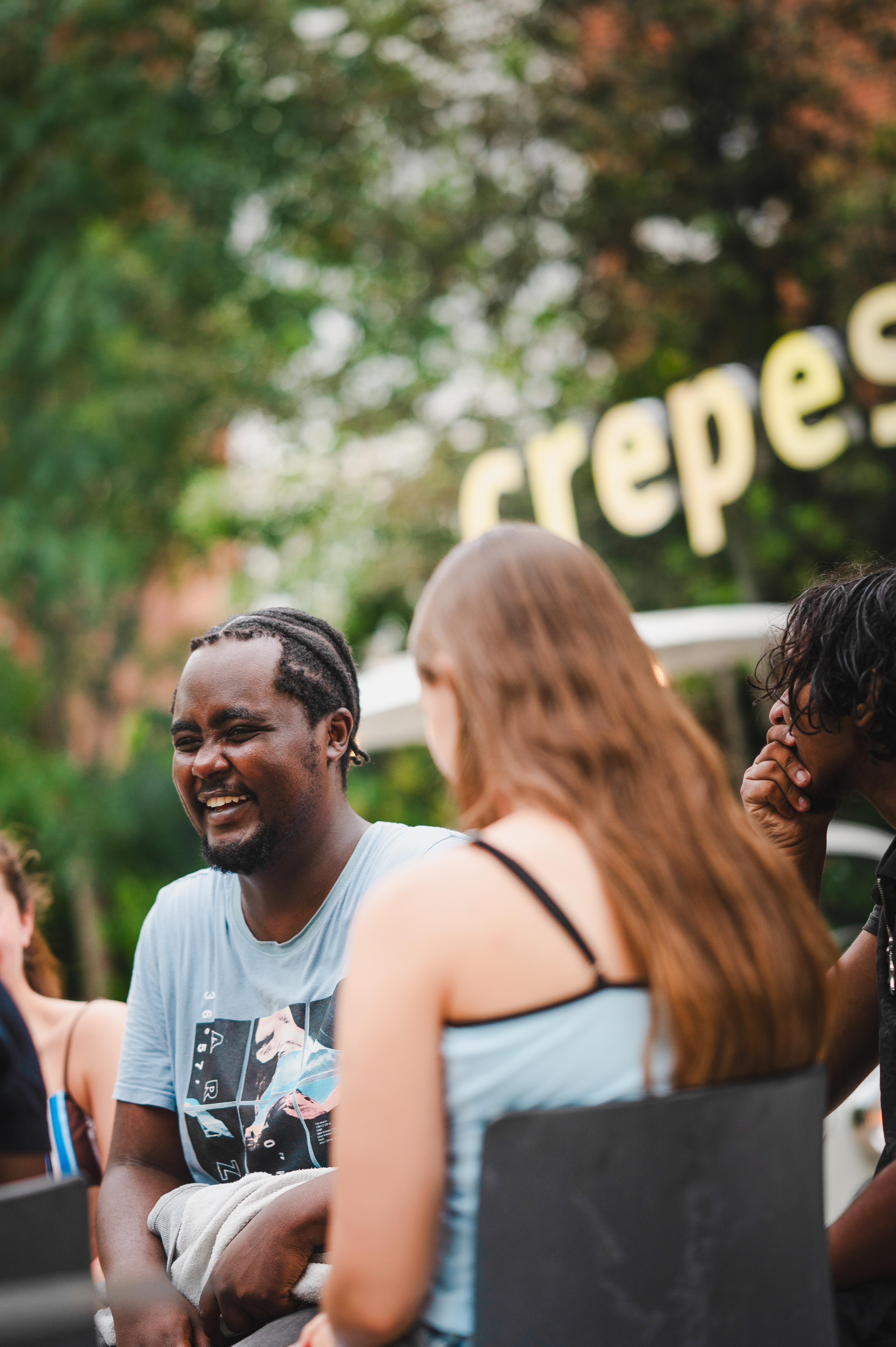 A man in a blue shirt smiles and talks with two women outdoors, with a “crepes” sign blurred in the background—an inviting scene that highlights the power of student social tips for making friends, even if you’re an introverted student.