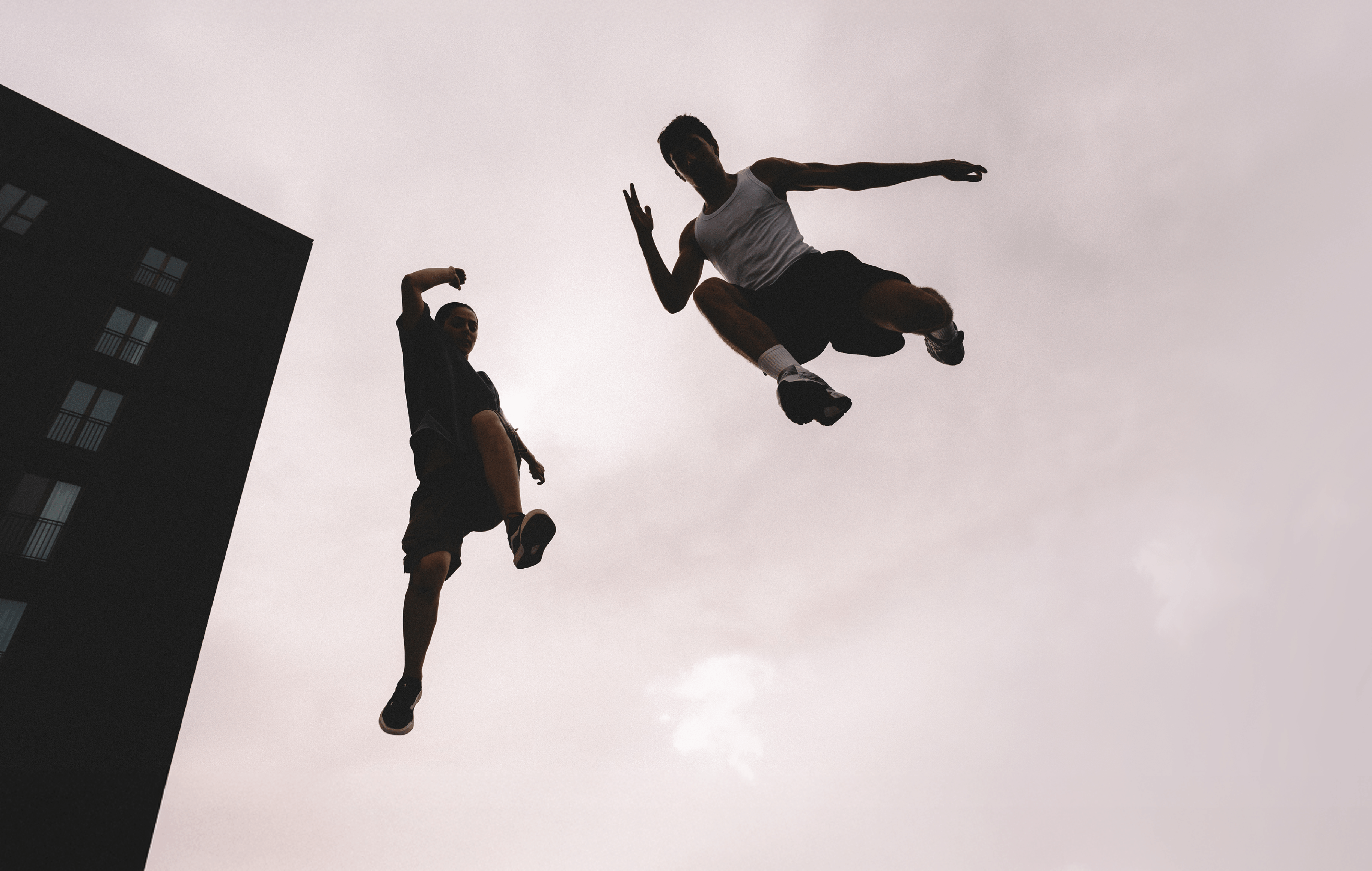Two people mid-air, jumping against a cloudy 2027 sky, with a dark building partially visible on the left side of the image.