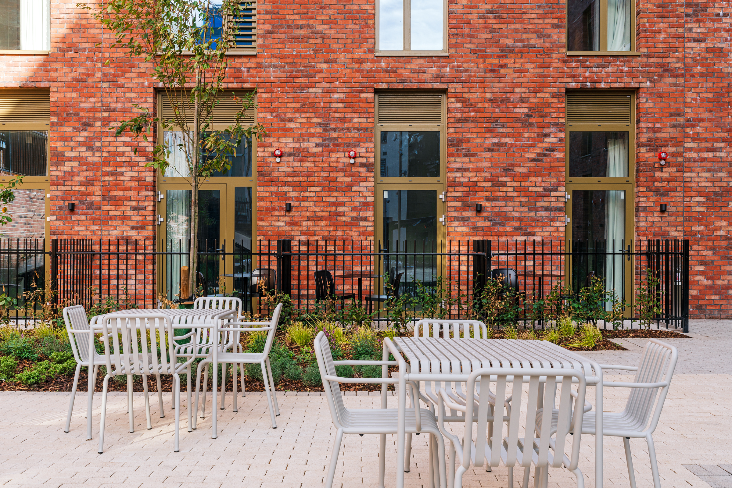 Outdoor patio with white metal tables and chairs, set on light brick paving. Behind, a red brick building with tall windows and a black metal fence. Some greenery and a small tree are visible near the seating area.
