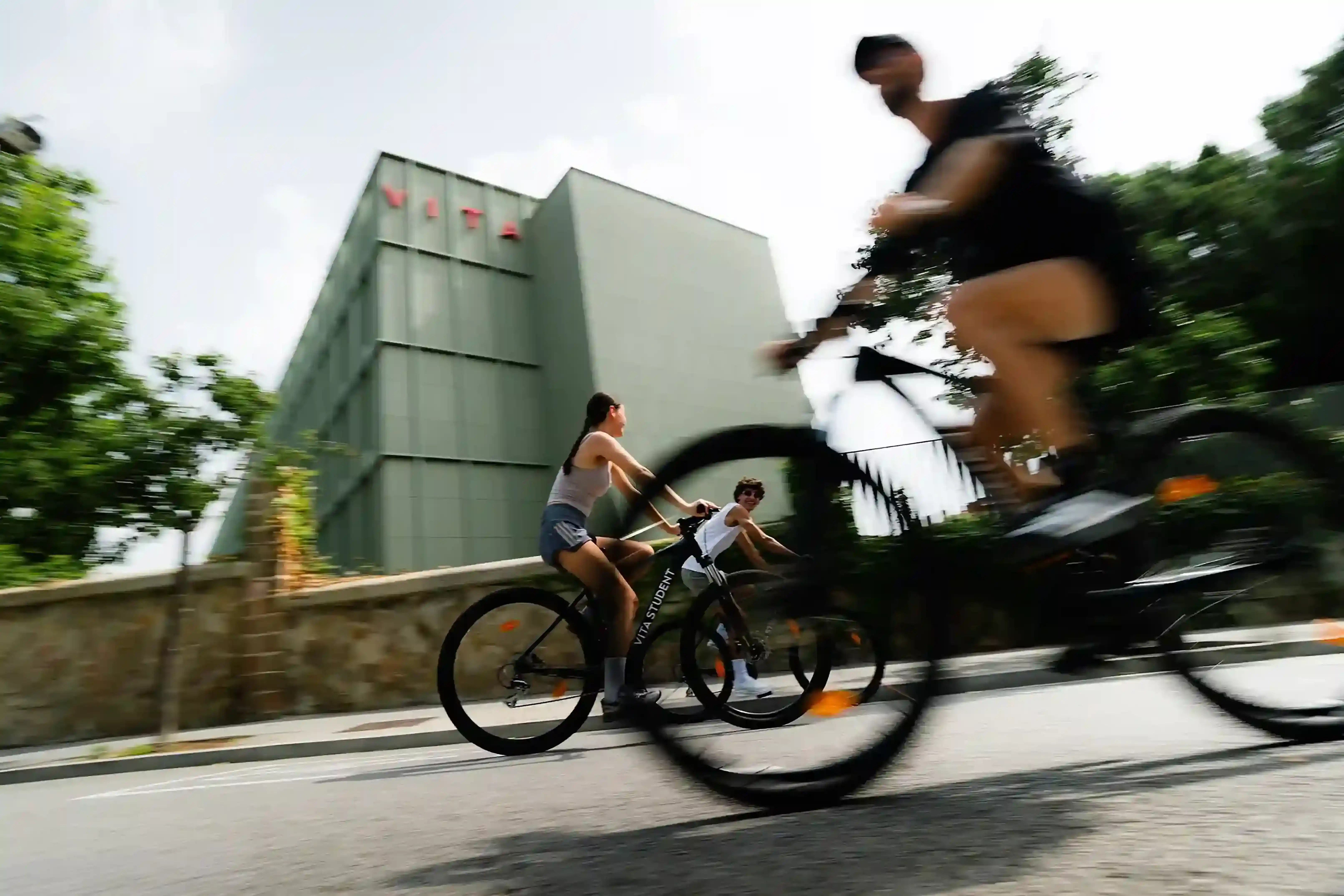 Three people ride bicycles on a sunlit street near a modern green building, possibly heading to an open day event; the closest cyclist appears blurred due to motion, while the background is clear.