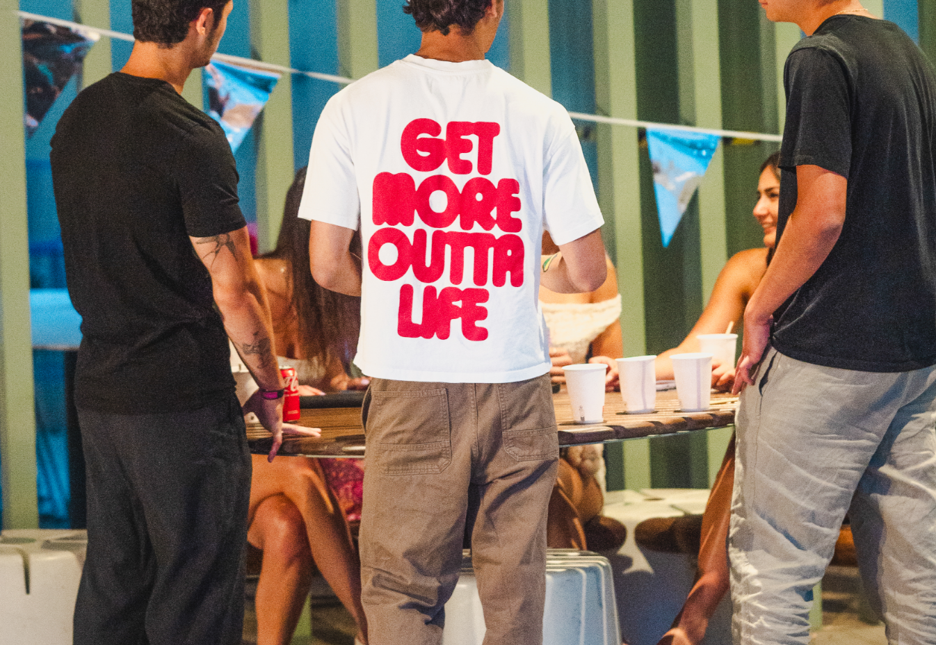 Four people stand near a table with cups and drinks, while two seated women smile. One person wears a shirt with large text reading “GET MORE OUTTA LIFE.” The scene, reminiscent of friends gathering to rebook plans or share discount offers, appears to be a casual indoor get-together.