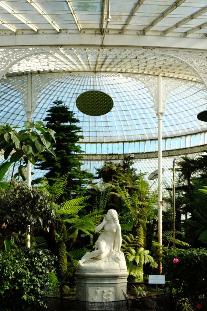 A marble statue surrounded by lush green plants inside a large glass conservatory with an ornate, domed roof and metal supports, this hidden gem is one of the top things to do in the West End—especially popular among students seeking inspiration and tranquility.