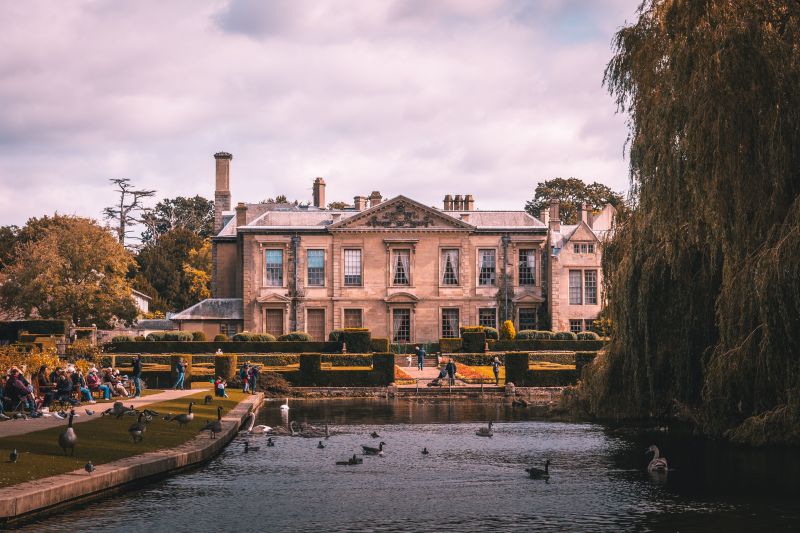 A large historic mansion sits behind manicured gardens and a pond with ducks and swans. People, including students on day trips from Coventry, are walking and sitting near the water under a cloudy sky.