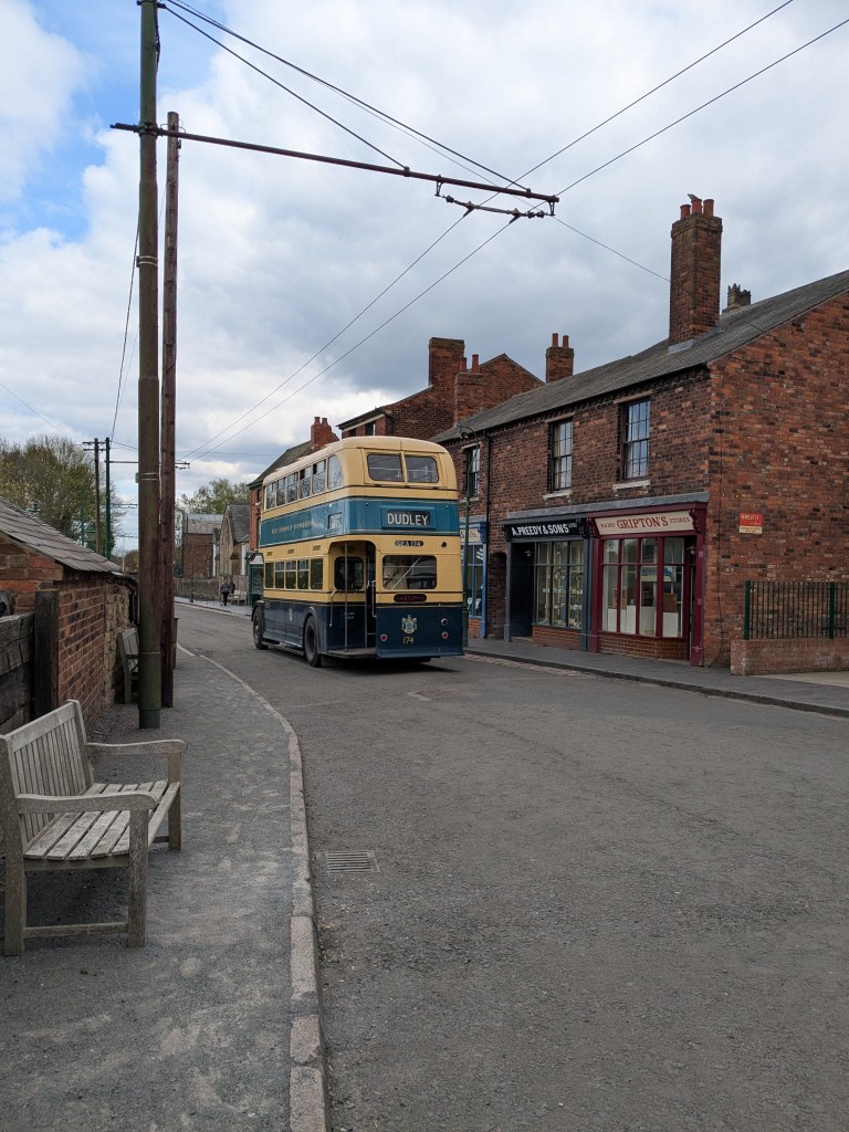 A vintage double-decker bus labeled “DUDLEY” drives down a quiet street in Coventry, lined with old brick buildings and shops under a partly cloudy sky. Perfect for day trips, the scene evokes memories of students waiting by the wooden bench that sits on the sidewalk in the foreground.