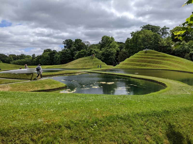 People, including students on day trips, walk on grassy mounds and paths winding around reflective ponds in a sculpted park landscape near Edinburgh, under a partly cloudy sky, with trees in the background.
