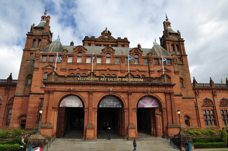 The Kelvingrove Art Gallery and Museum in Glasgow, Scotland, features a grand red sandstone facade, arched entrances, and Scottish flags on display under a partly cloudy sky. Located in the vibrant West End, it’s a popular spot for students seeking interesting things to do in the city.