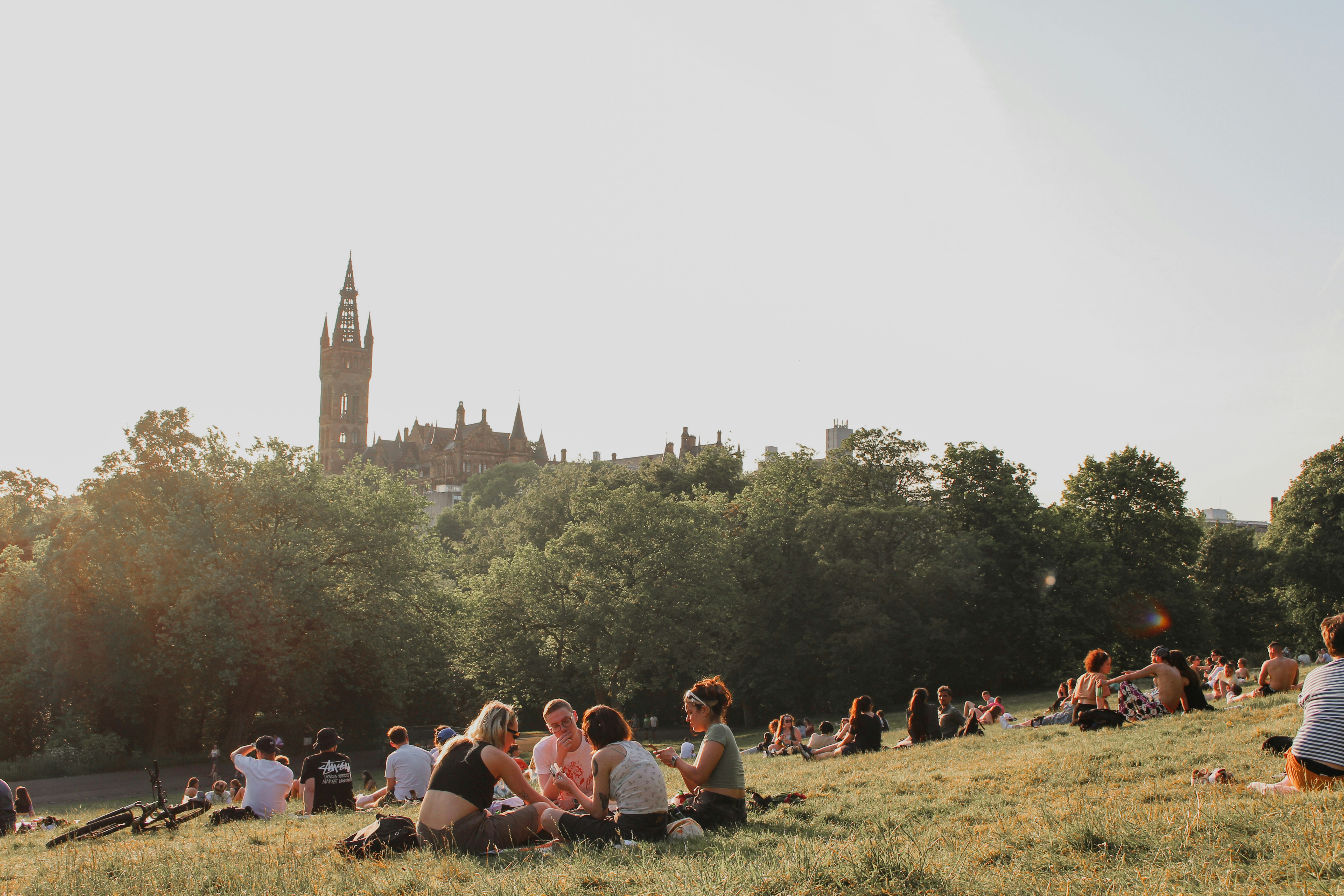 Groups of people, including students, sit and relax on a grassy hill in a park with trees and a large, ornate building with a clock tower in the background under a clear sky—one of the many popular things to do in the West End.