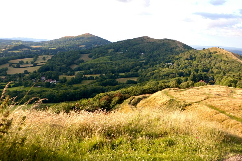 A scenic landscape of rolling green hills and fields under a partly cloudy sky, with tall grass in the foreground and wooded areas and scattered houses in the distance—perfect for students seeking peaceful day trips outside Coventry.