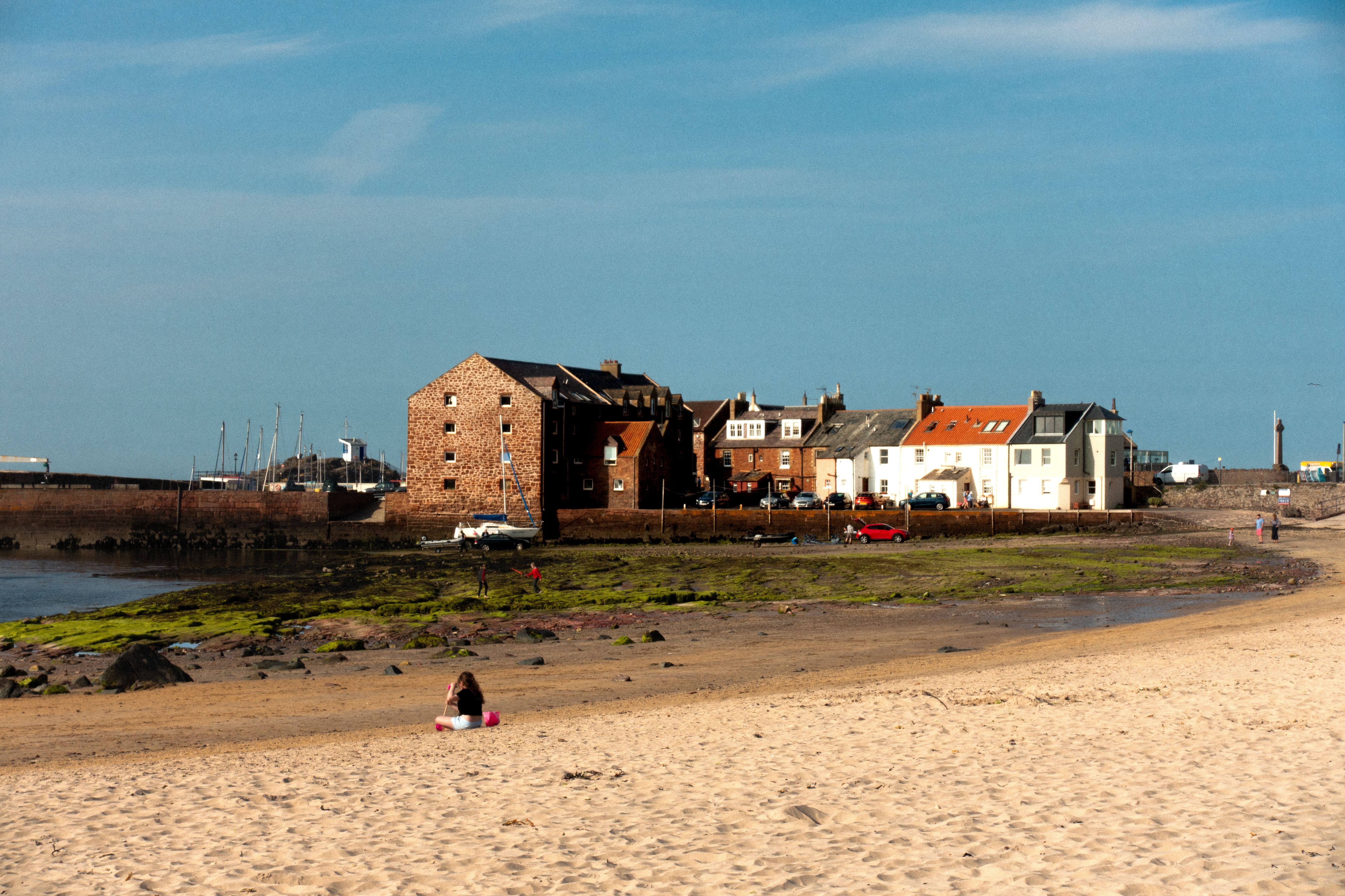 A sandy beach with one person sitting on the shore, colorful houses and brick buildings in the background, and a bright blue sky overhead—an ideal spot for students seeking relaxing day trips from Edinburgh.